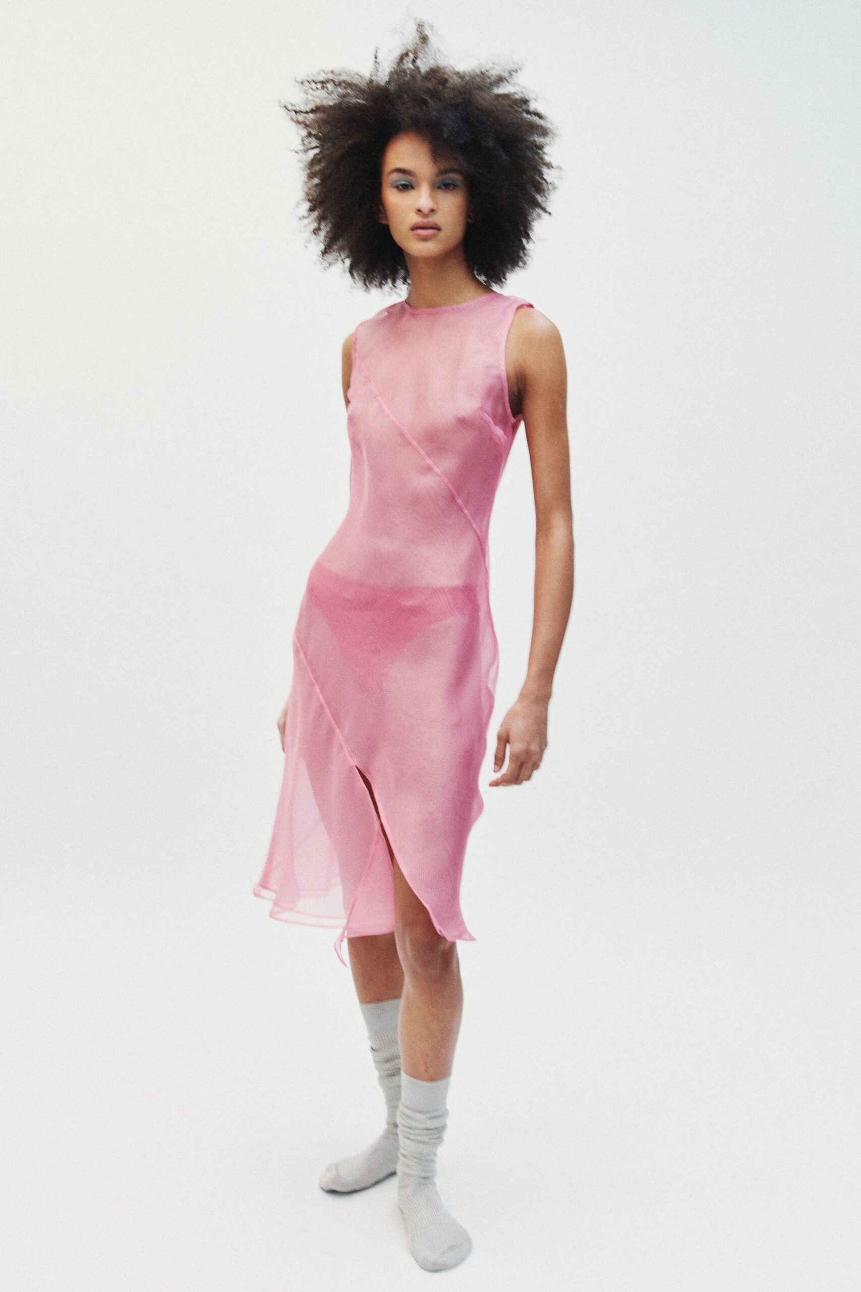 Sheer pink slip dress with curved seaming and a wavering front slit. Deeper rose briefs visible beneath the translucent skirt. Pale grey ribbed socks slouched softly at the ankle. Morning in a borrowed studio, heater off, someone still taping marks on the floor.