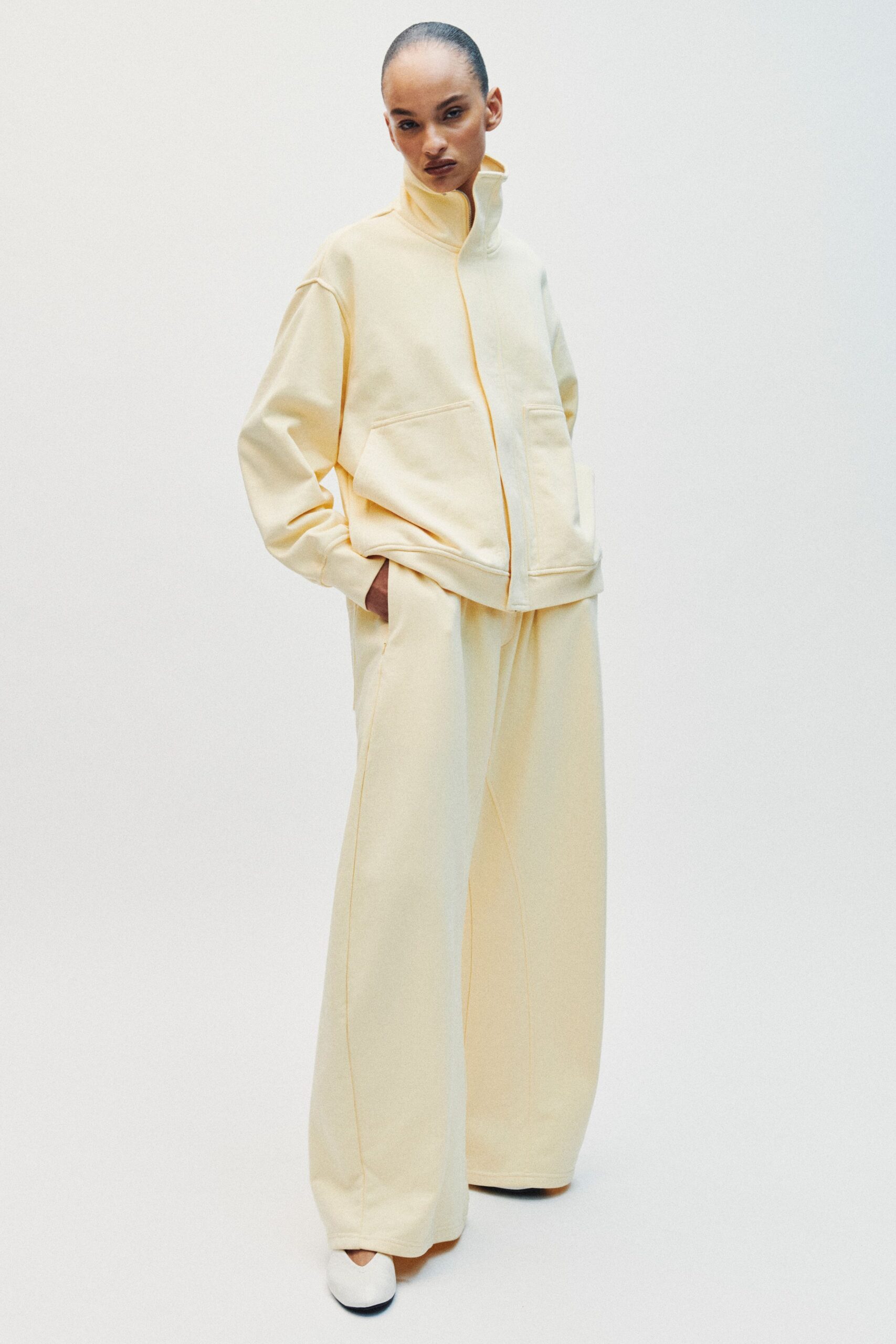 Buttercream brushed jersey zip jacket with a tall funnel neck. Matching wide-leg trousers with pressed front seams and a soft break. White rounded flats, just peeking from the sweeping hem. First flight out, airport windows still blue with morning rain.