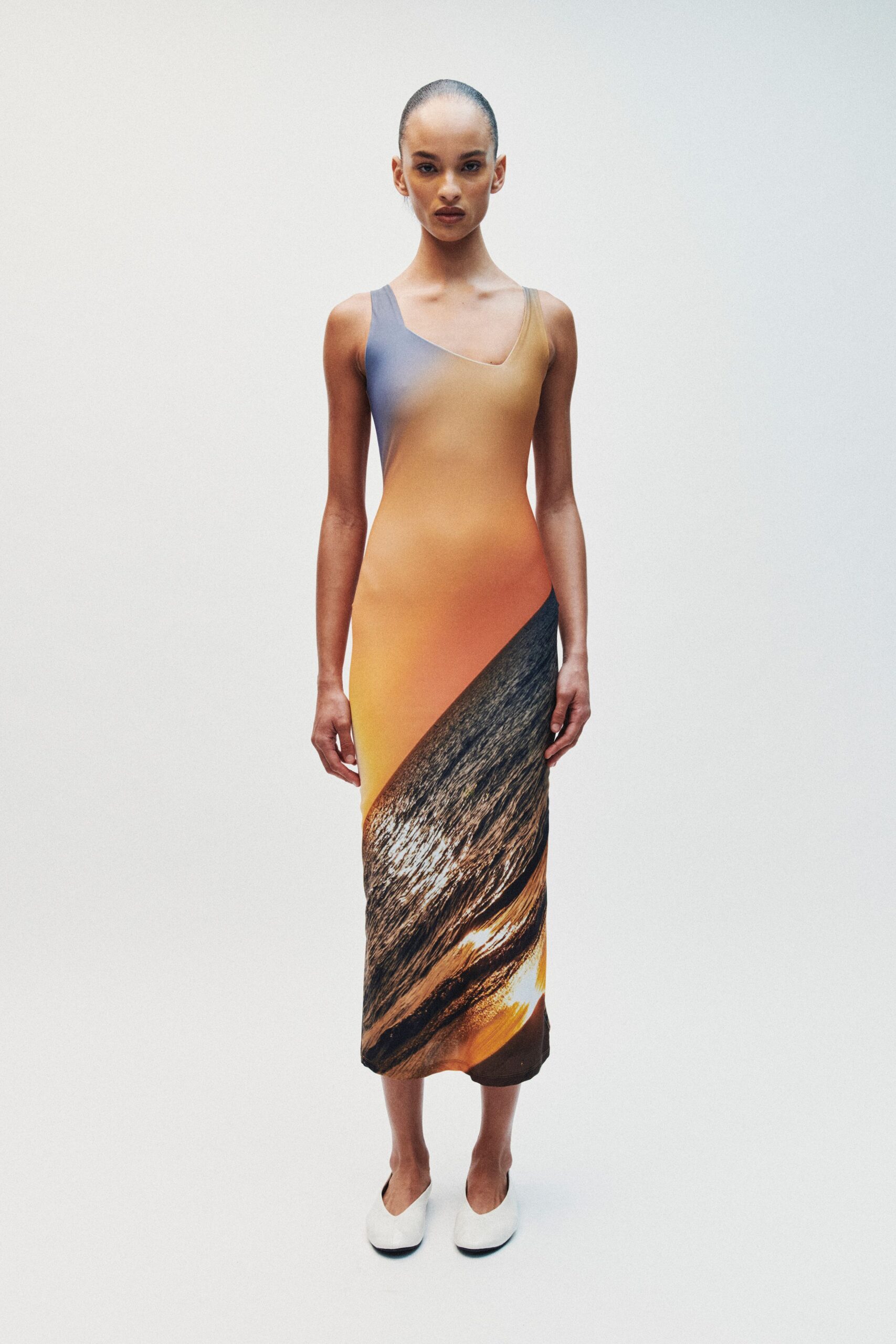 Asymmetric tank dress in glossy stretch jersey, sunset gradient breaking into a dark brushstroke hem. White square-toe flats with a deep vamp curve. Museum atrium at dusk, the skylight still holding heat.