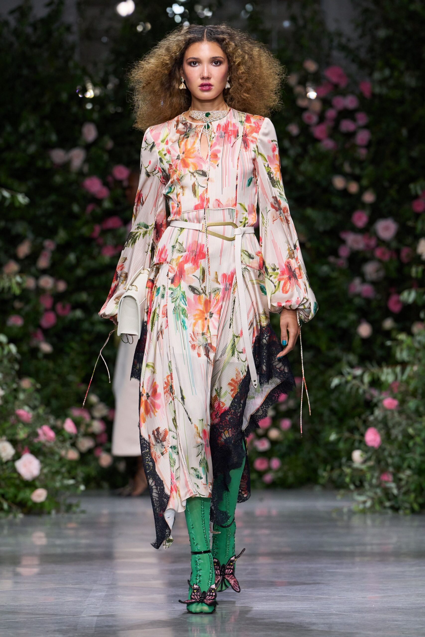 Cream micro-pleated dress washed with coral florals and black lace drifts. Ivory leather tie belt looping through a pale gold buckle. Emerald knee boots with butterfly appliques opening over the toes. Tiny bell-shaped drops. Green cabochon at the throat. Walking the long path to a conservatory wedding after rain.