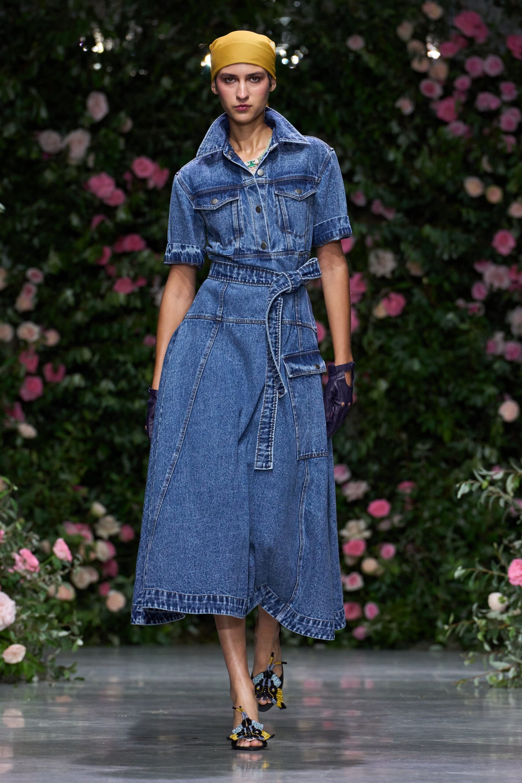 Mustard stretch headscarf pulled smooth and close over the crown. Acid-wash denim shirtdress with a popped collar and self-belt knot. Navy fingerless gloves cut away across the knuckles. Black sandals topped with beaded moth wings and yellow tassels. Walking the rose path after rain, hem picking up greenhouse damp.