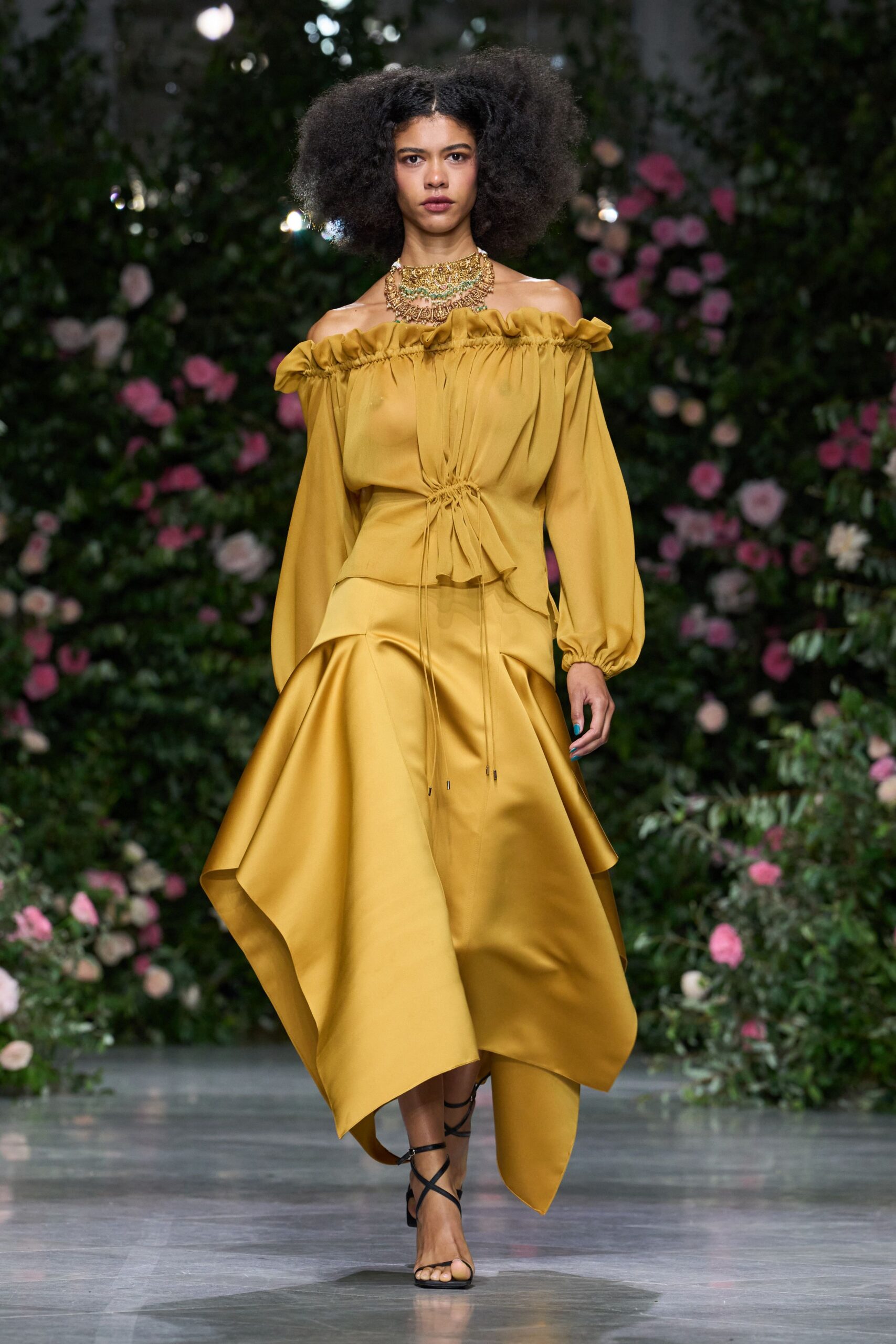 Layered gold filigree collar with pale green stones at the throat. Mustard satin dress, ruffled off-the-shoulder neckline and drawstring waist with tipped cords. Black wrap sandals with thin straps crossing the bare foot. Crossing a conservatory aisle while the orchids fog up the glass.