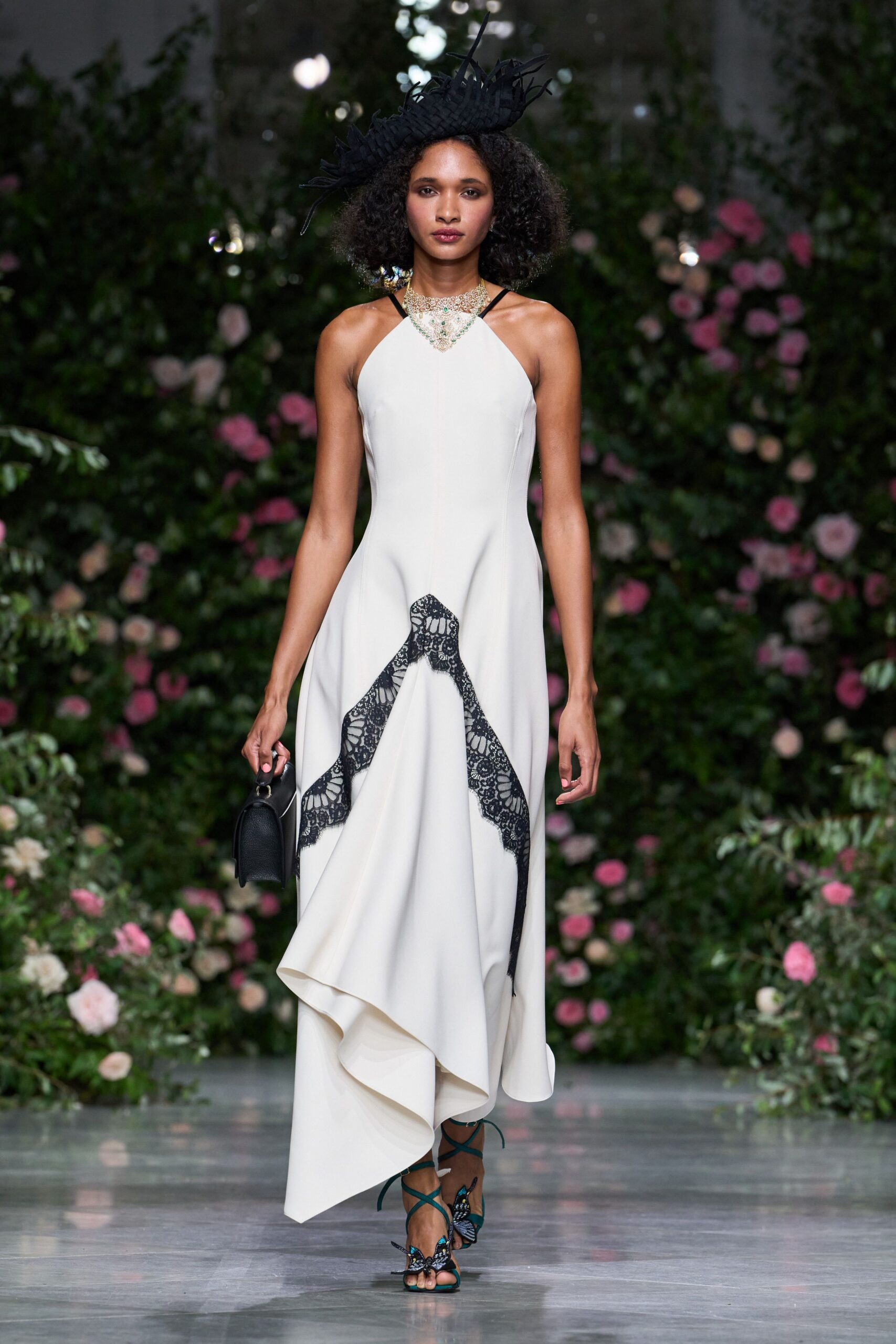 Black woven hat bristling with narrow cut strips. Crystal bib necklace with pale green stones at the center. Ivory halter gown with black lace appliqued in a climbing curve. Black pebbled top-handle bag. Teal lace-up sandals with butterfly wings over the toes. Garden wedding after midnight rain, stepping carefully off the grass.