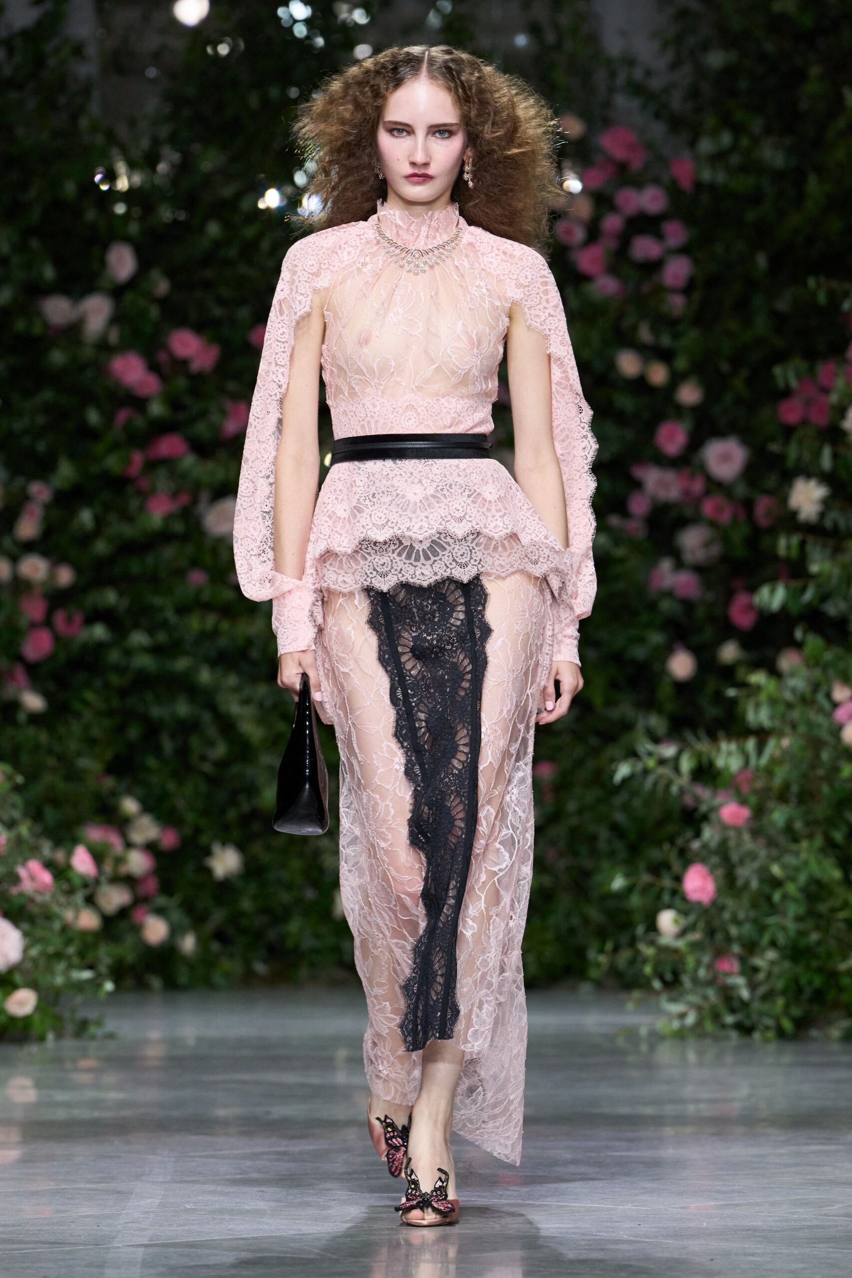 Blush floral lace dress with a sheer yoke and winged slit sleeves. Narrow black satin belt cinching the scalloped peplum. Black lace inset falling from the hip like a misplaced slip. Glossy black folded clutch. Open-toe mules topped with pink butterfly appliqués. Garden wedding reception, champagne gone warm under the marquee.