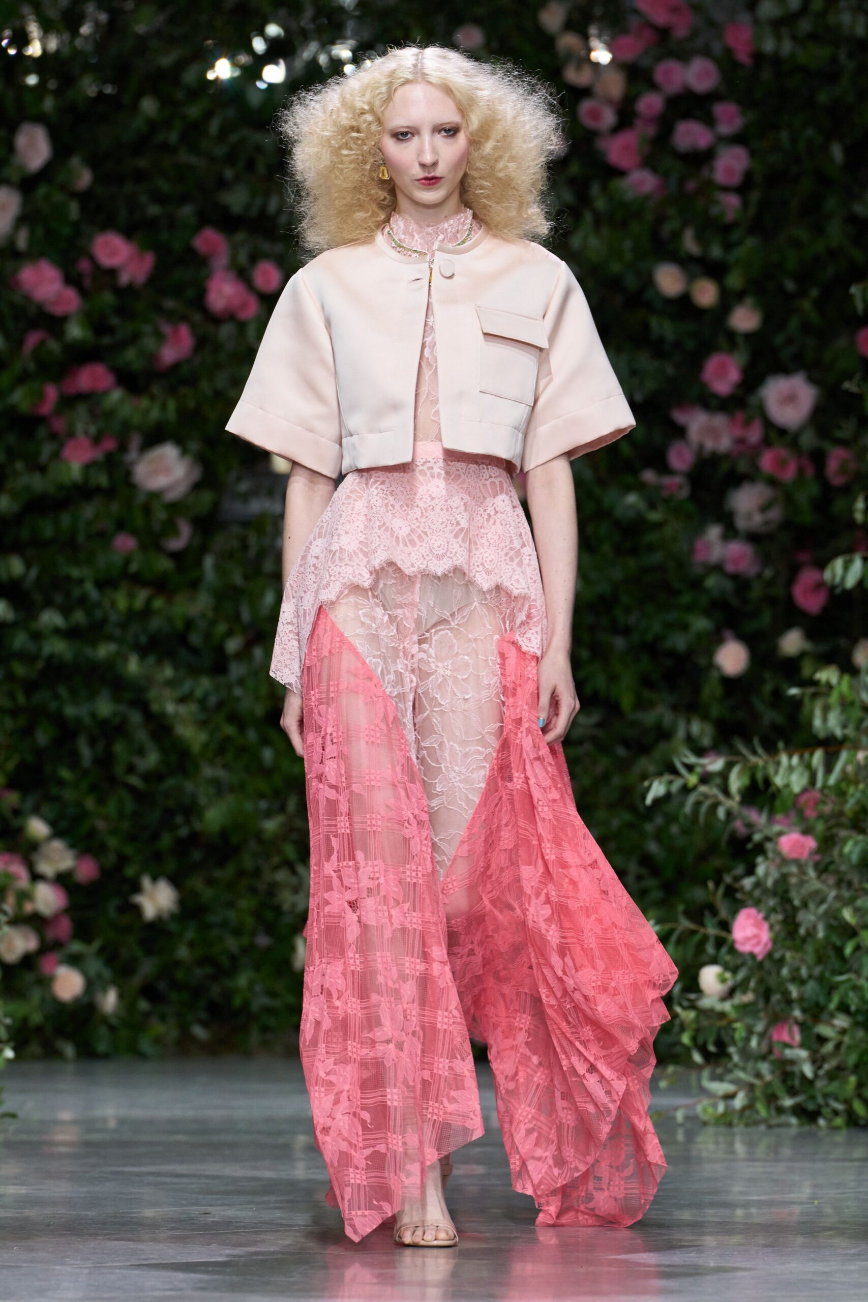 Blush satin cropped jacket with broad turned cuffs and one flap pocket. Sheer shell-pink lace rising to a ruffled mock neck. High-low lace skirt, pale floral panel opening into hot-coral sides. Barely-there clear strap sandals. Garden wedding reception, cake cut and the grass already damp.