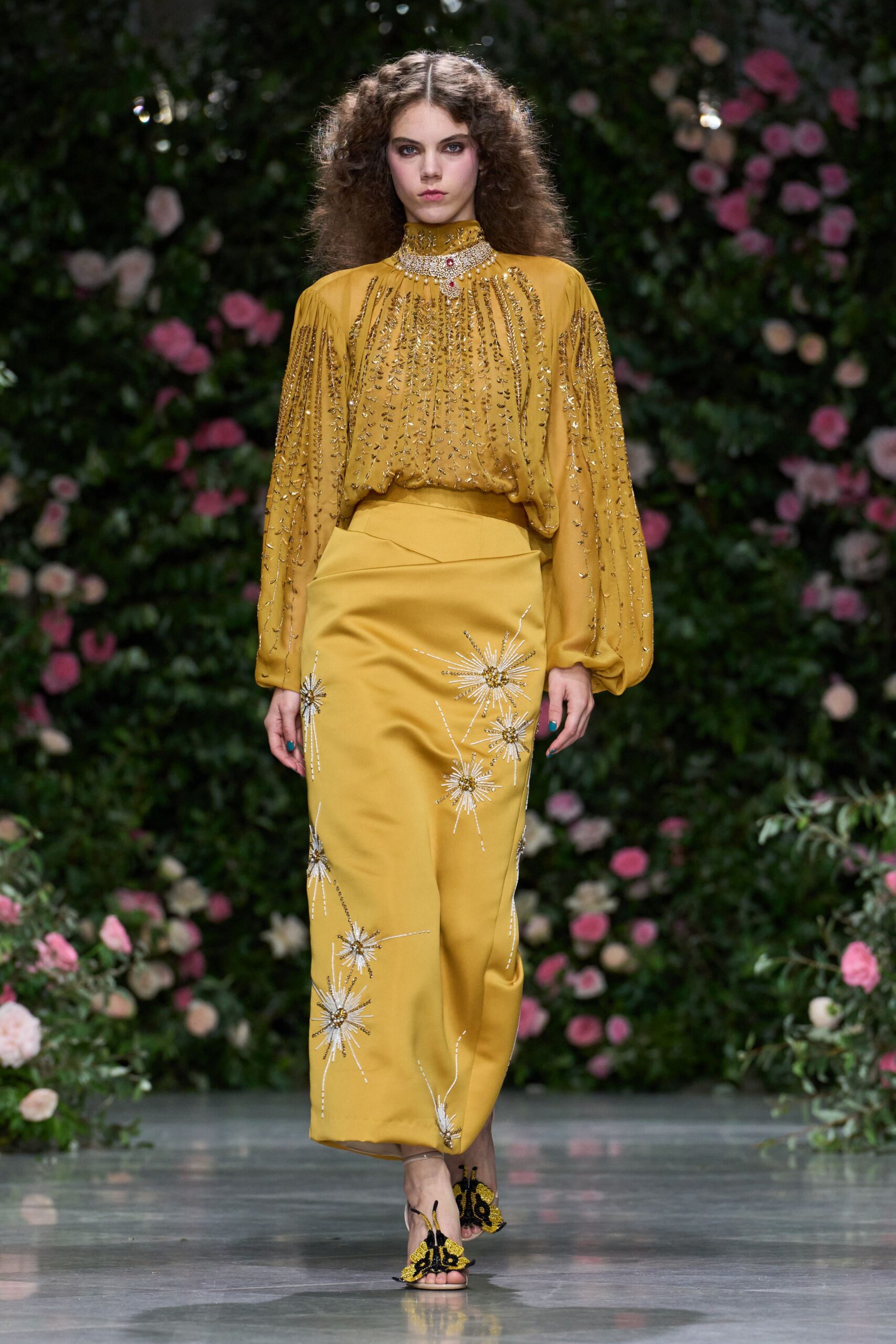 Ochre georgette blouse draped loose, sleeves streaked with dangling gold beading. Crystal collar necklace with a red stone drop at the throat. Matching column skirt scattered with silver starburst beading, one burst at the hem. Black-and-gold butterfly sandals. Garden wedding at the hour when the champagne has gone warm.
