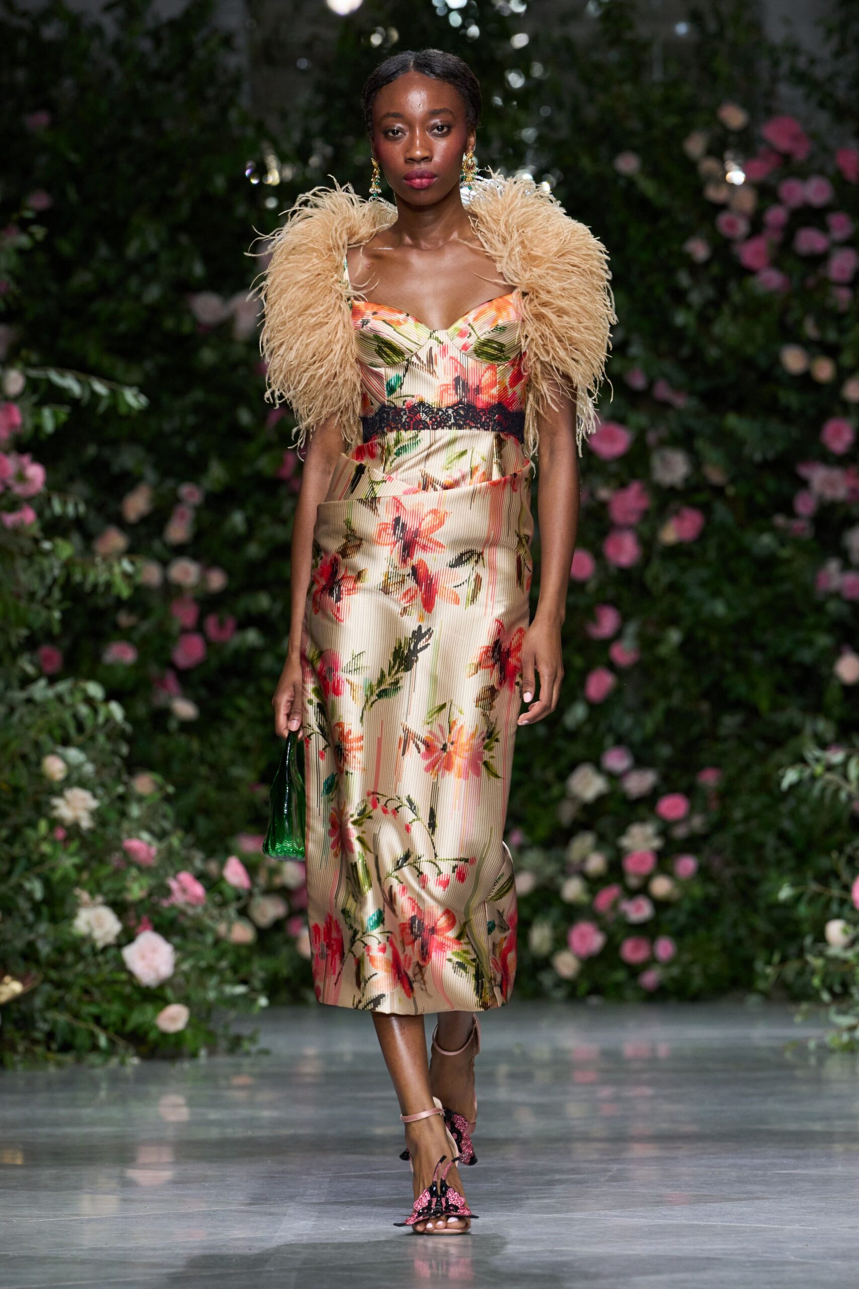 Champagne pleated satin dress with painterly flowers and black lace underbust. Straw-beige ostrich-feather stole swelling high around both shoulders. Small emerald beaded pouch. Pink floral sandals with black bow tufts at the toe. Garden wedding, heat still rising off the stone after sunset.