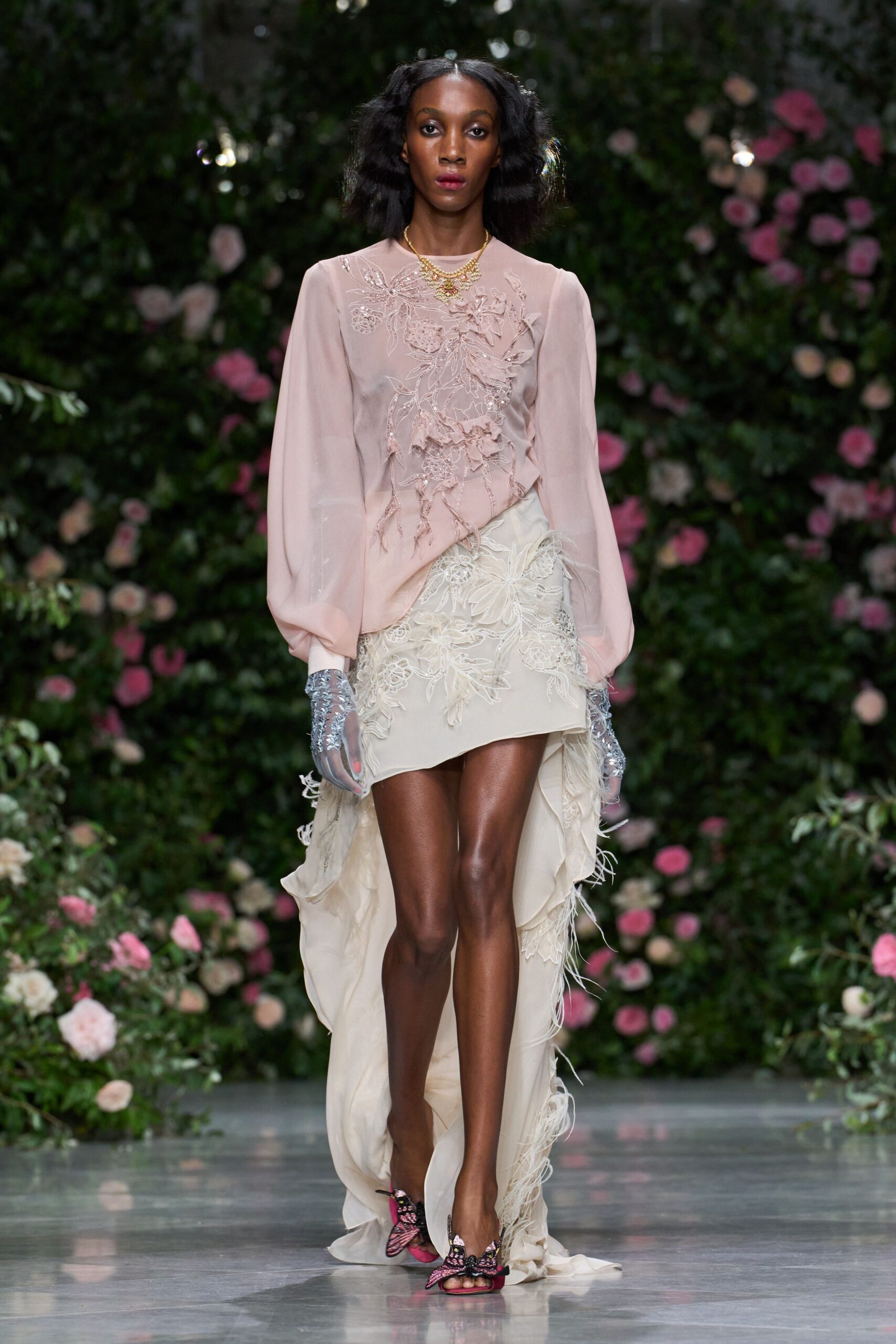 Blush georgette blouse with trailing floral appliqué across the chest. Ivory chiffon skirt cut high-low, feathered lace spilling from the hem. Sheer gloves crusted in pale silver beading. Hot-pink sandals topped with beaded butterfly wings. Garden wedding, cake already cut and the grass damp again.