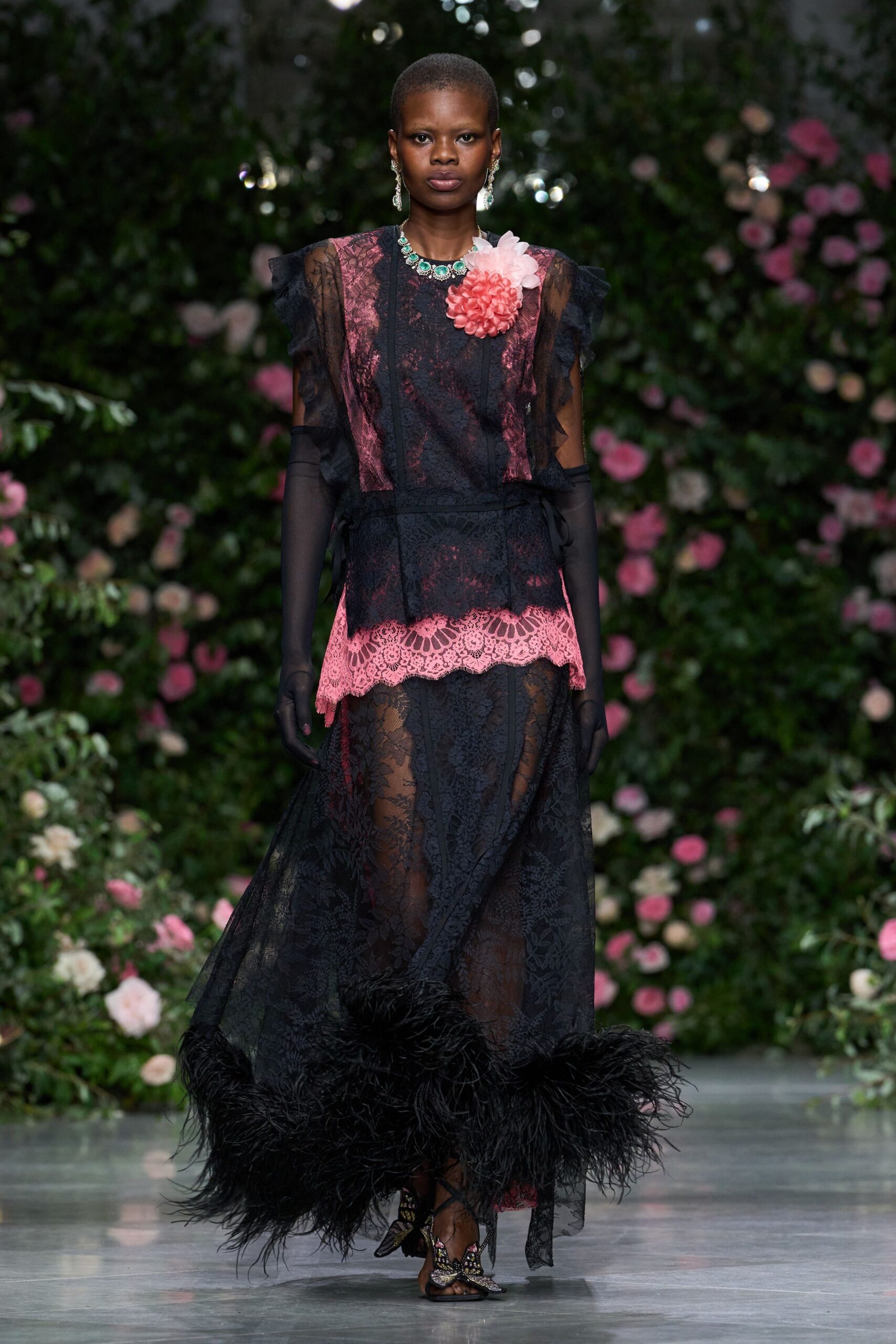 Turquoise stones in a bright silver collar. Black floral lace dress over pink underlay, feathered at the sweeping hem. Sheer black opera gloves ruched softly at the upper arm. Butterfly sandals glittering with beadwork at the toe. Garden wedding gone humid, roses dropping petals onto the path.