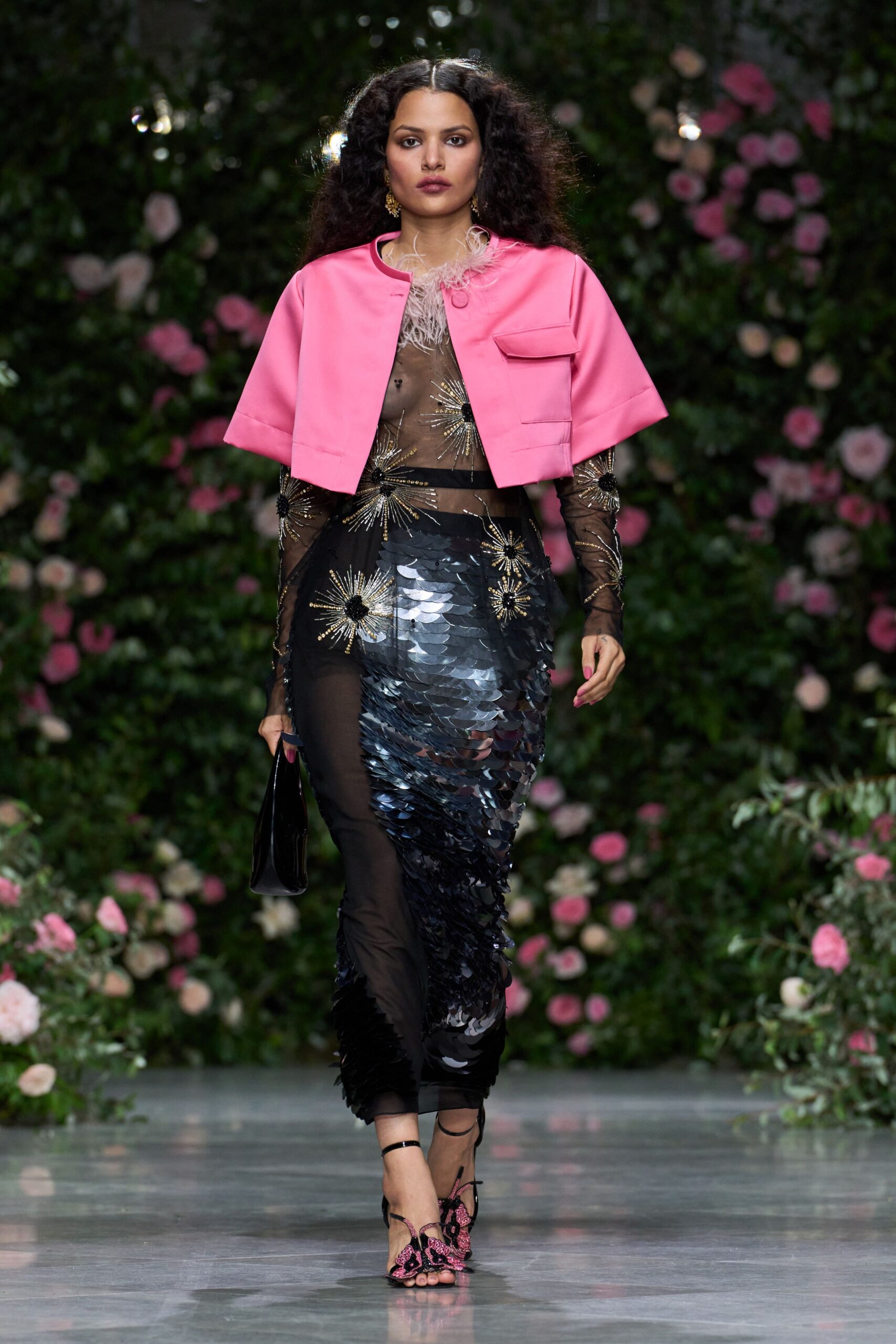 Bubblegum satin cape jacket, cropped and boxy with a single flap pocket. Sheer black mesh dress spangled with gold starbursts and oversized paillettes. Glossy black clutch tucked flat in the hand. Ankle-strap sandals with pink beaded butterfly fronts. Crossing a hotel conservatory after midnight with petals stuck to your hem.