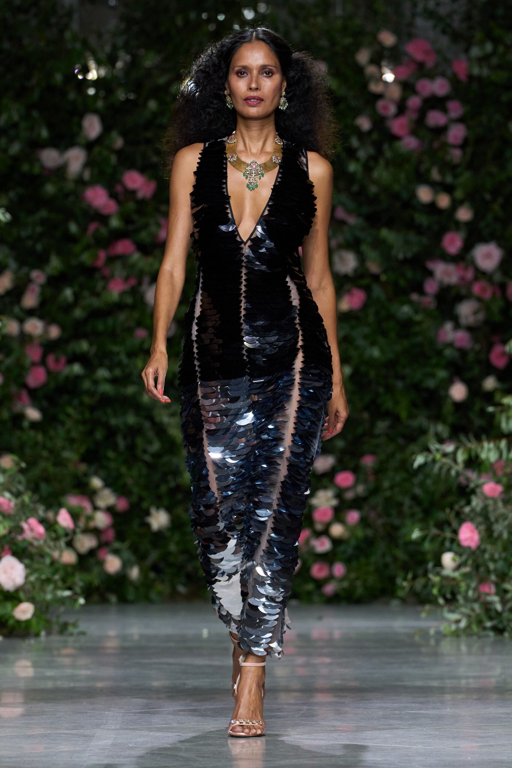 Rigid gold collar set with pink and emerald stones. Plunging sheer column dress covered in oversized black paillettes, one spine-straight seam. Nude ankle-strap sandals with a jeweled toe band. Walking out to the terrace after midnight, roses still holding the day's heat.