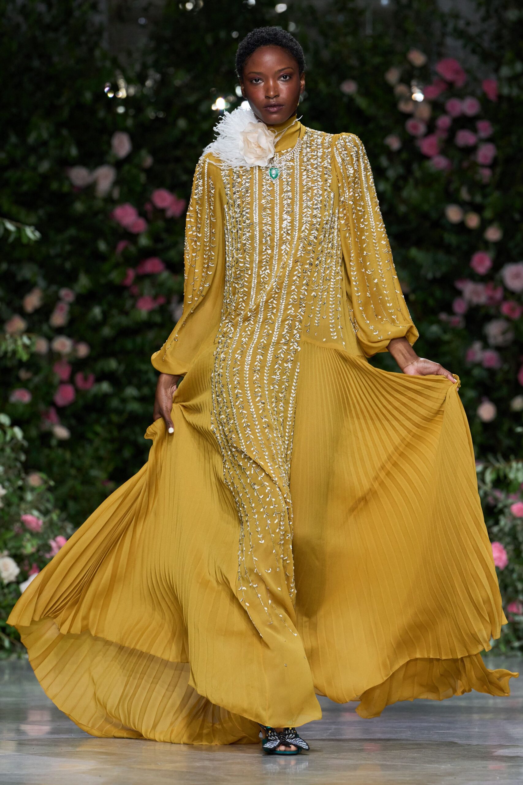 Mustard chiffon gown with knife-pleated skirt and silver vine beading. White feather burst pinned with an oversized fabric camellia. Green teardrop stone at the throat. Beaded bow sandals, toes just visible. Garden wedding after rain, crossing the lawn before dusk.