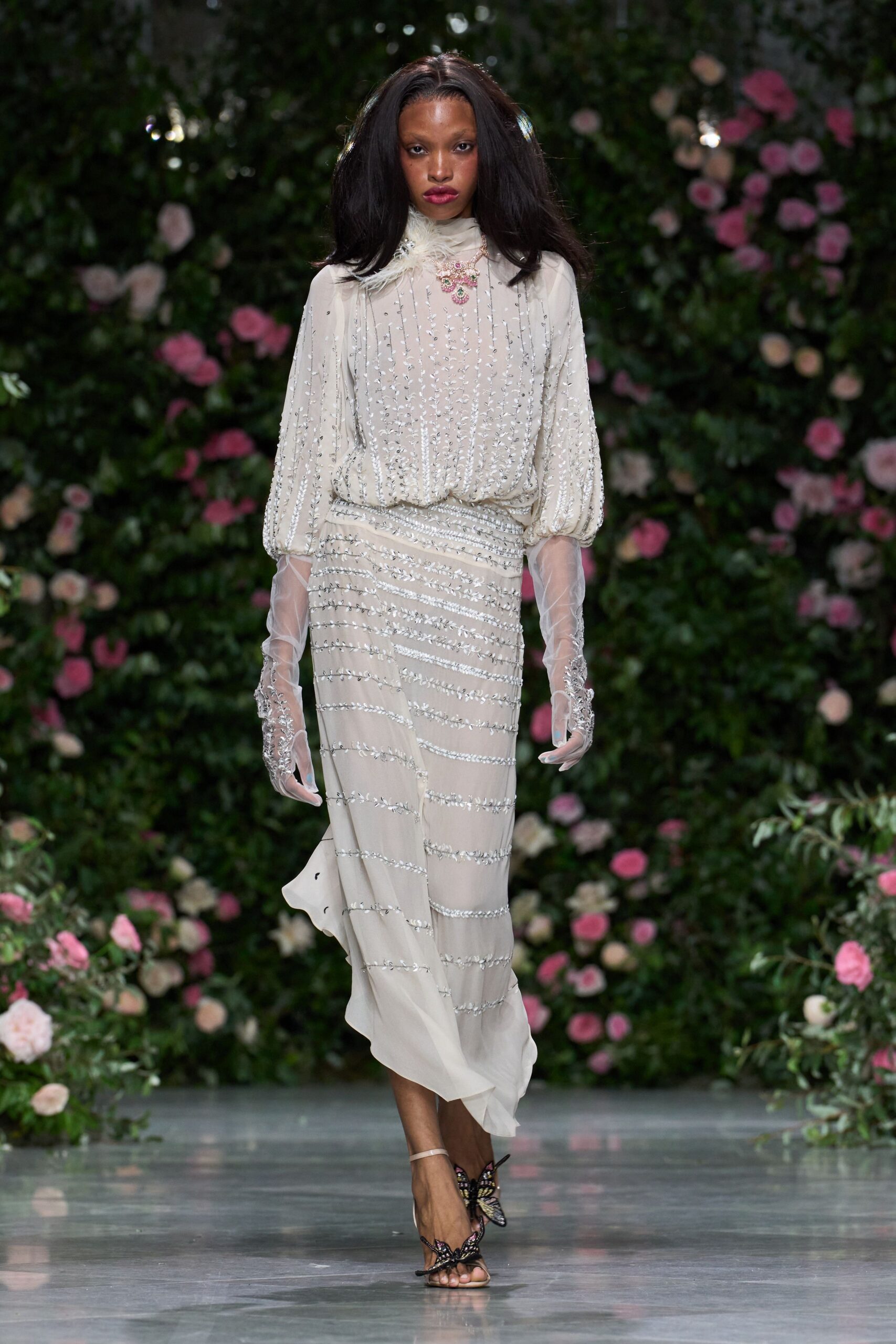 Ivory chiffon dress with silver vine beading and a low wrapped hip. Sheer tulle opera gloves, beadwork clustering at the wrists. Pink jeweled pendant over a small white feather tuft. Butterfly sandals with beaded wings perched over the toes. Crossing a conservatory aisle while the wedding guests are still finding seats.