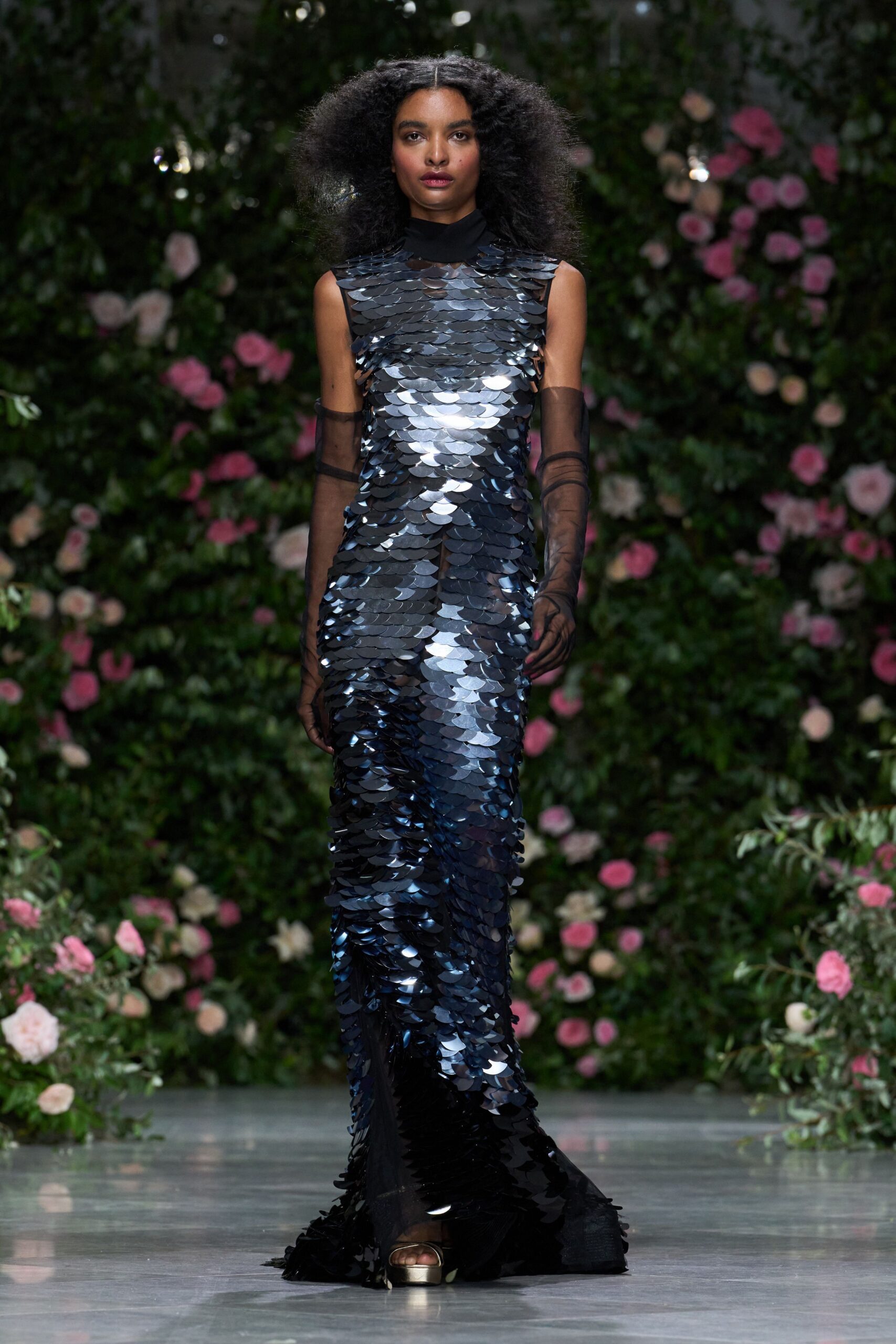 Sleeveless black column gown covered in oversized paillette discs, sheer at the throat. Smoke tulle opera gloves, one finger seam slightly twisting. Pale metallic platforms peeking beneath the hem. Crossing a conservatory aisle after dark, roses gone sweet and damp.
