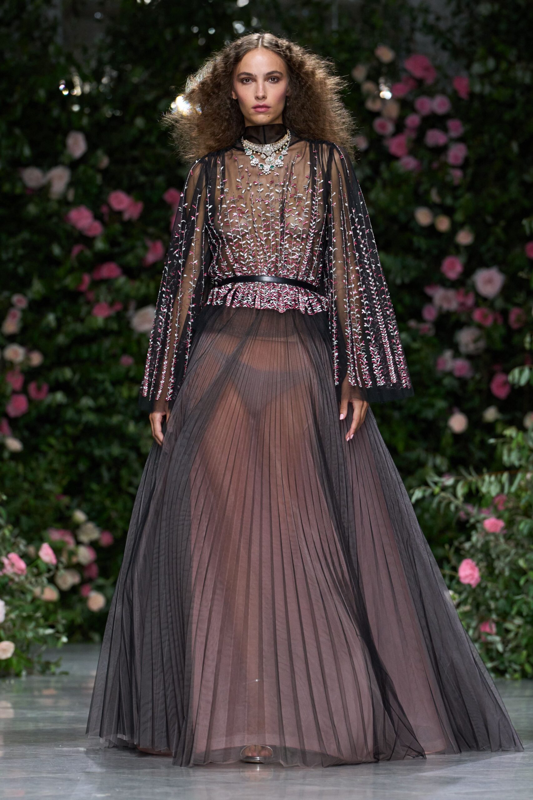 Tiered crystal necklace with mint stones clustered tight at the throat. Sheer black tulle gown, pink-and-silver sprig embroidery above a knife-pleated skirt. Slim black waistband cutting the transparency at the smallest point. Garden wedding after midnight, damp grass catching at the hem.