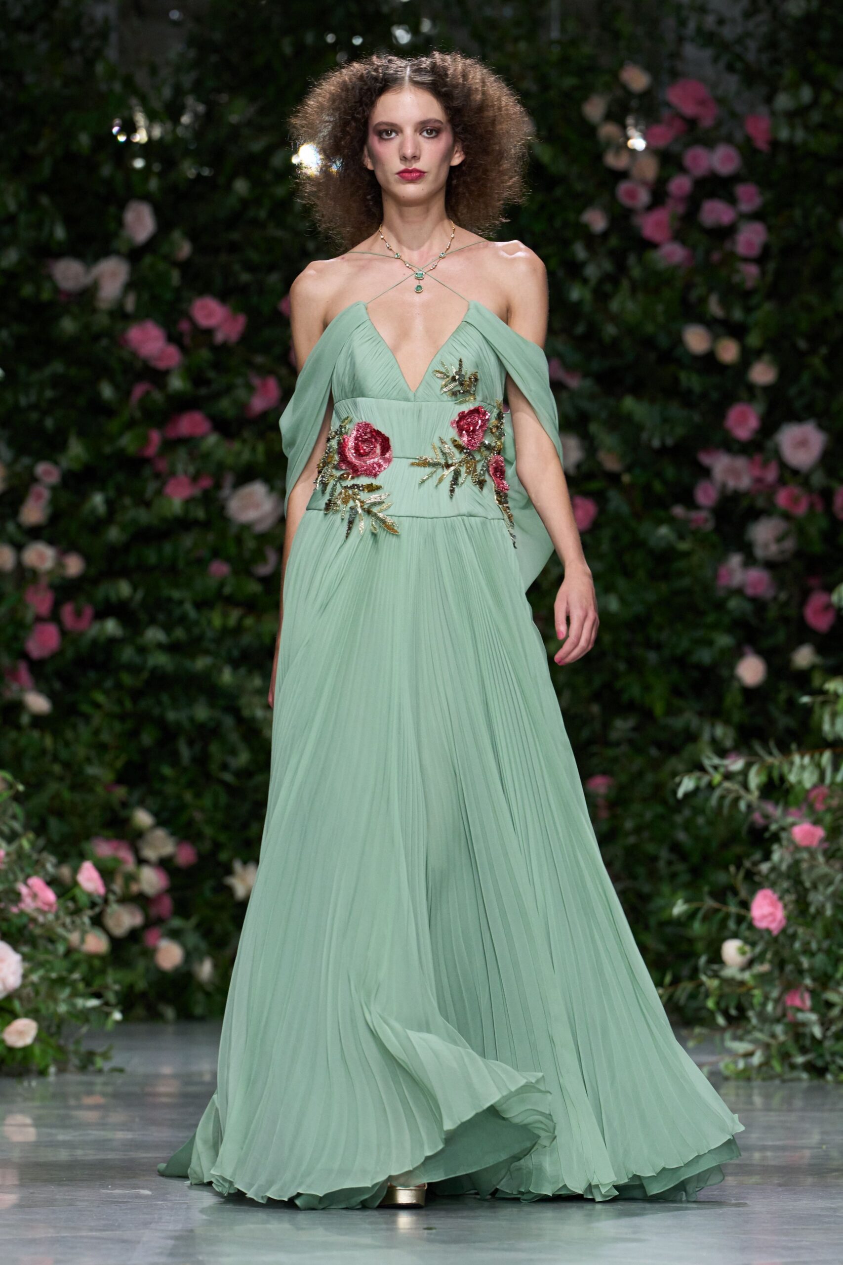 Fine gold chain necklace crossed at the collarbones with green stones. Seafoam chiffon gown, knife-pleated through the skirt with sequined rose appliques and draped off-shoulder panels. Pale metallic platforms peeking from the hem. Crossing the lawn after a wedding, grass still wet underfoot.