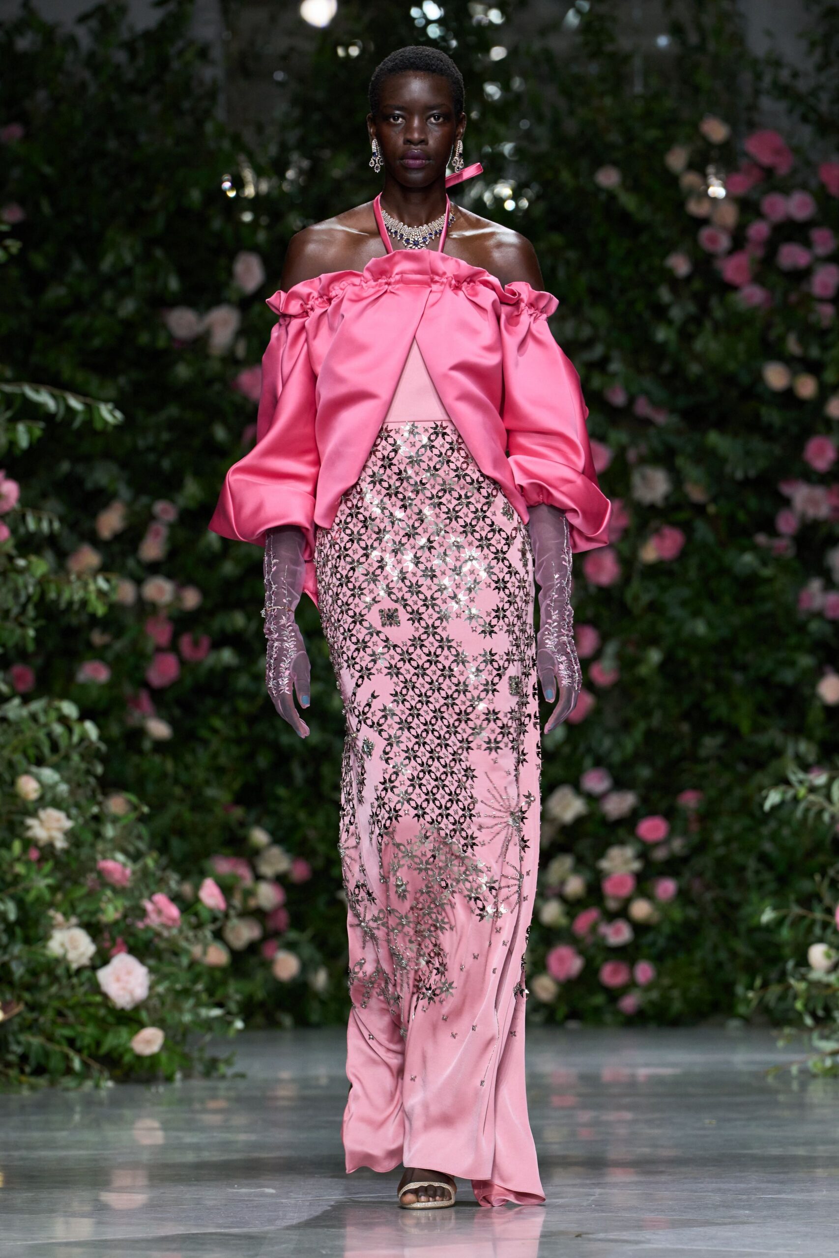 Bubblegum satin cape top, ruched off the shoulder with swollen sleeves. Blush column gown with silver beadwork and a narrow midriff cutout. Sheer mauve gloves with beaded vines climbing the forearm. Crystal-trim slide sandals. Crossing a hotel conservatory after the wedding guests have gone upstairs.