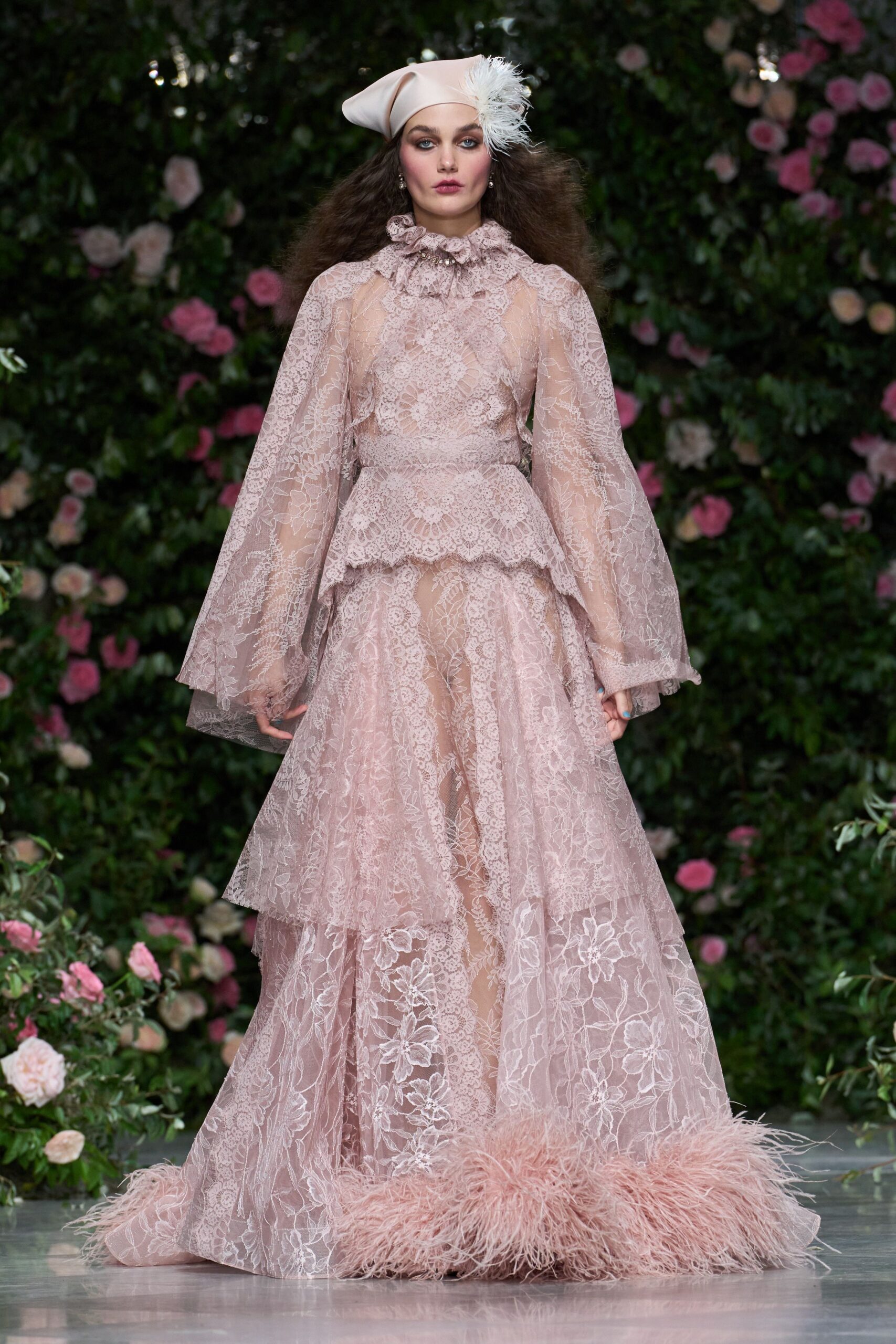 Blush satin cap tipped forward, pinned with a white feather burst. Powder-pink lace gown with ruffled collar, sheer bell sleeves, and ostrich-feather hem. Small drop pearls at each ear. Garden wedding after the rain, gravel still dark underfoot.