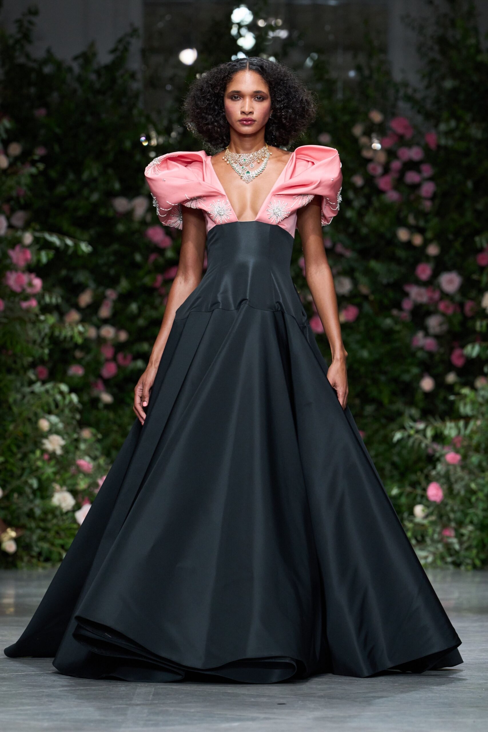 Tiered crystal bib necklace with pale green stones at the center. Black satin ballgown with a plunged bodice and shell-pink sculpted sleeves, beaded starbursts scattered at the bust. Garden wedding after midnight, the roses still holding the day's heat.