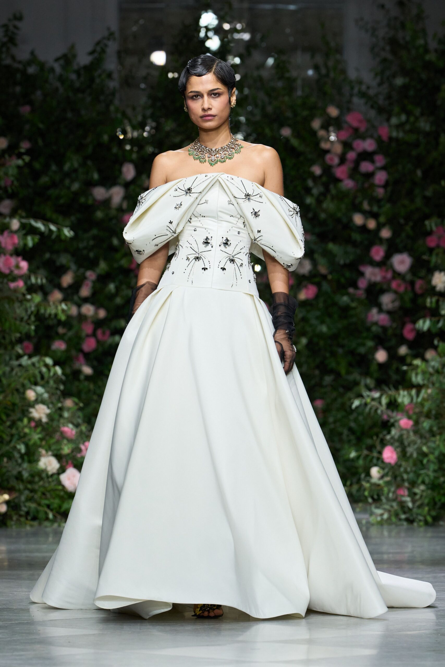 Diamond collar with a fringe of pale green pear drops. Ivory strapless ballgown with folded off-shoulder sleeves and black starburst beading. Sheer black tulle gloves ruched to the forearm. Black floral sandals peeking from beneath the hem. Walking through a conservatory wedding after the cake has already been cut.