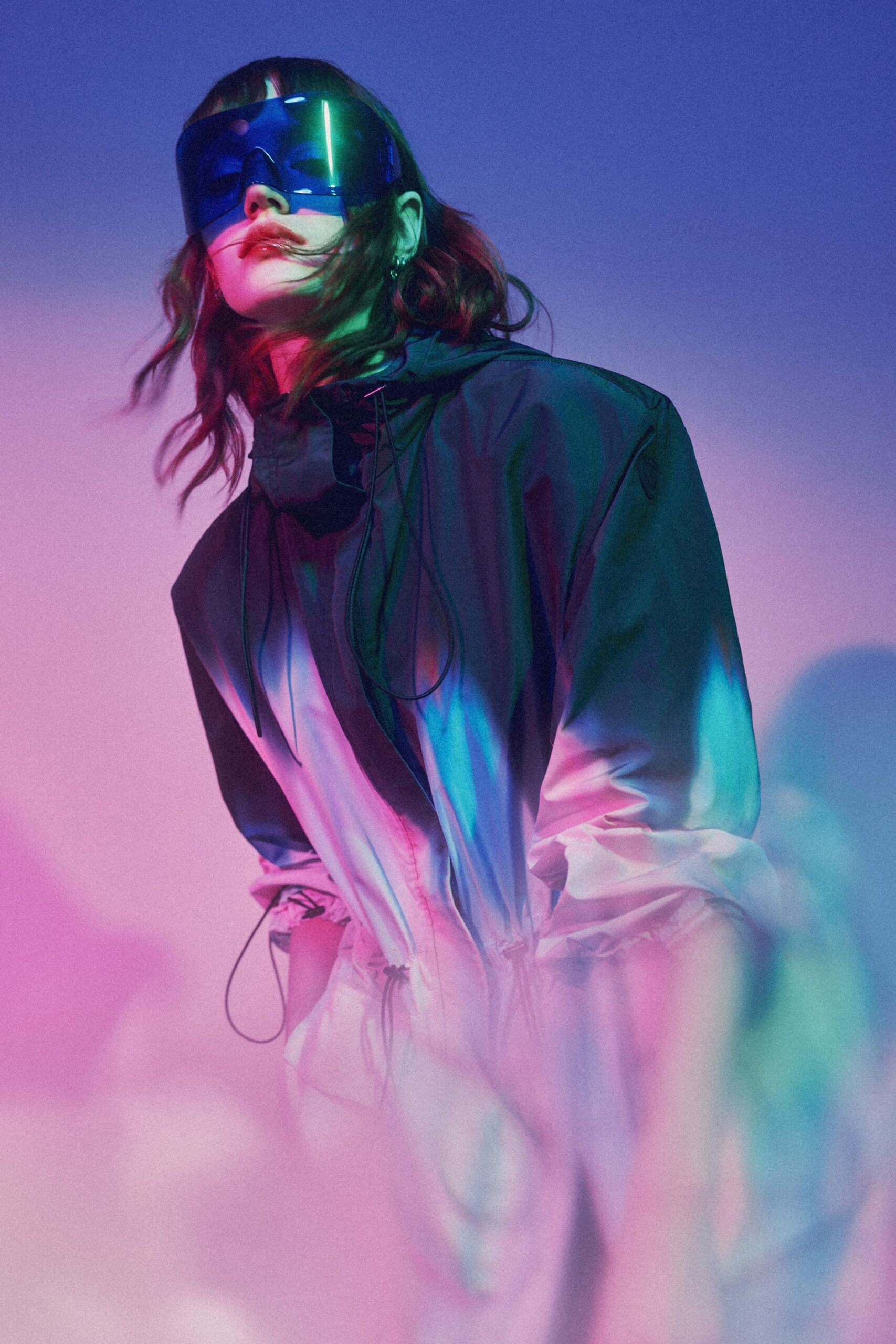 Oversized cobalt shield visor with a single uninterrupted curved lens. Black nylon anorak fading to chalk white, bungee cords dangling loose at the throat. Crossing a rooftop in colored fog before the storm siren test.