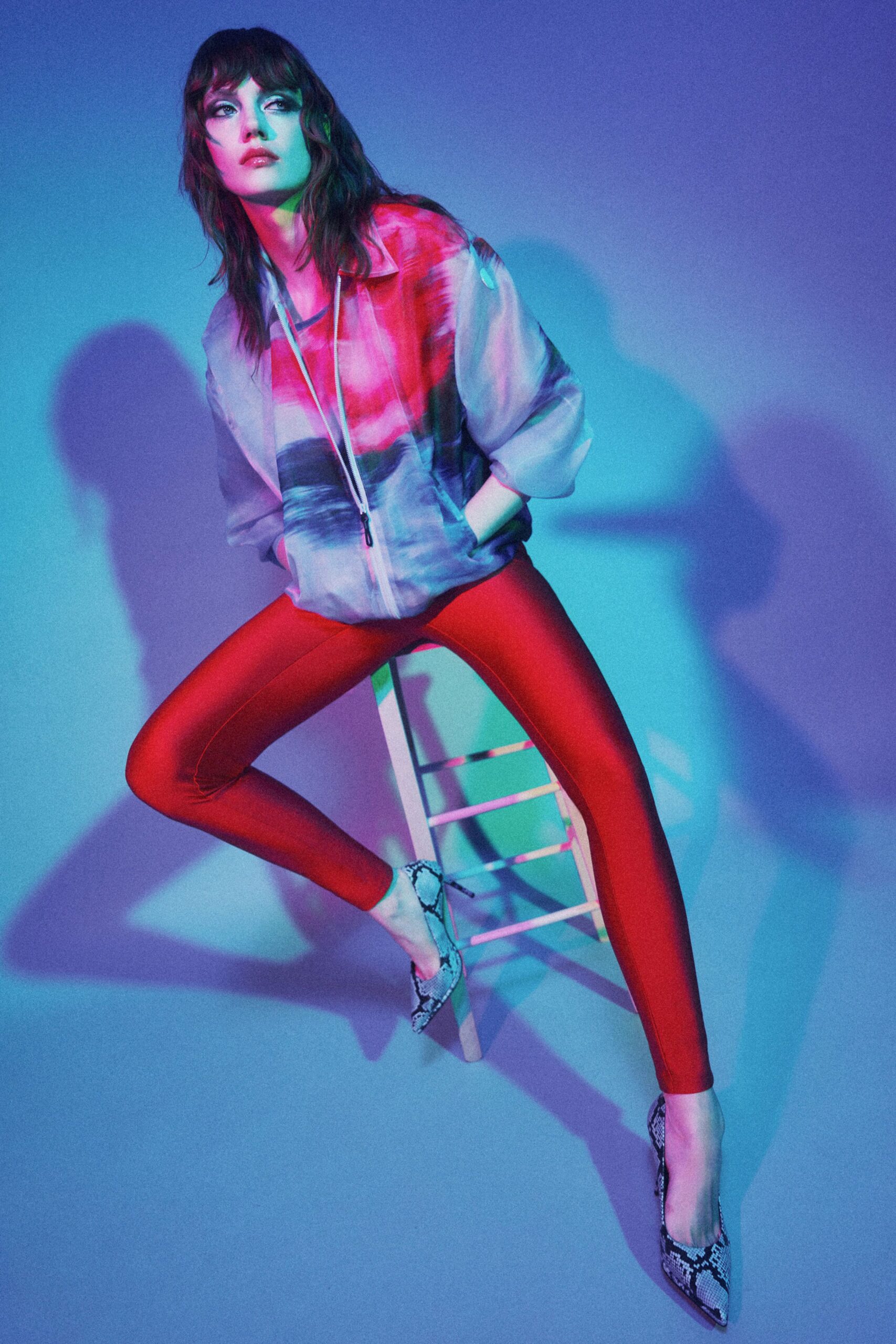 Sheer zip-front hoodie washed in magenta and slate, black pull dangling low. Tomato-red stretch leggings with a clean center seam. Gray python pumps with narrow pointed toes. Under club spill light, perched on a stool before midnight tips.