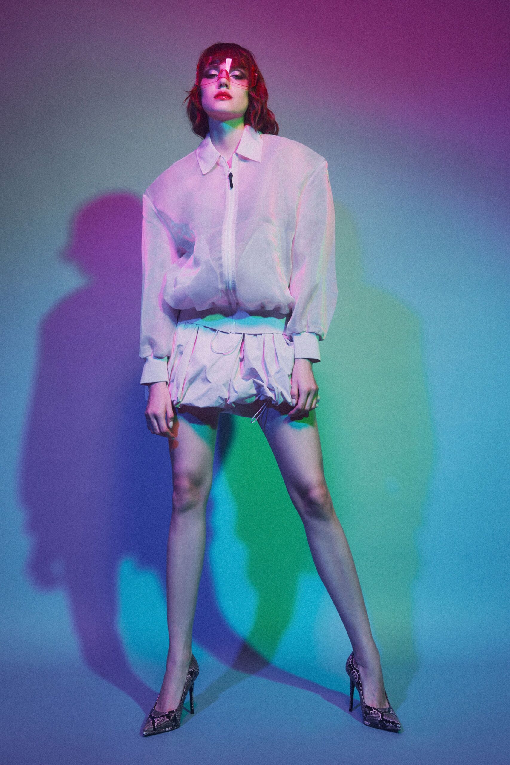Rose-tinted acrylic visor with a narrow white strip at center. Sheer white zip-front blouson, elastic hem ballooning above the waist. Matching paper-light shorts, ruched into a bubble hem with dangling cords. Python pumps with razor-point toes. Under club spill light, waiting for the elevator to open on the roof.