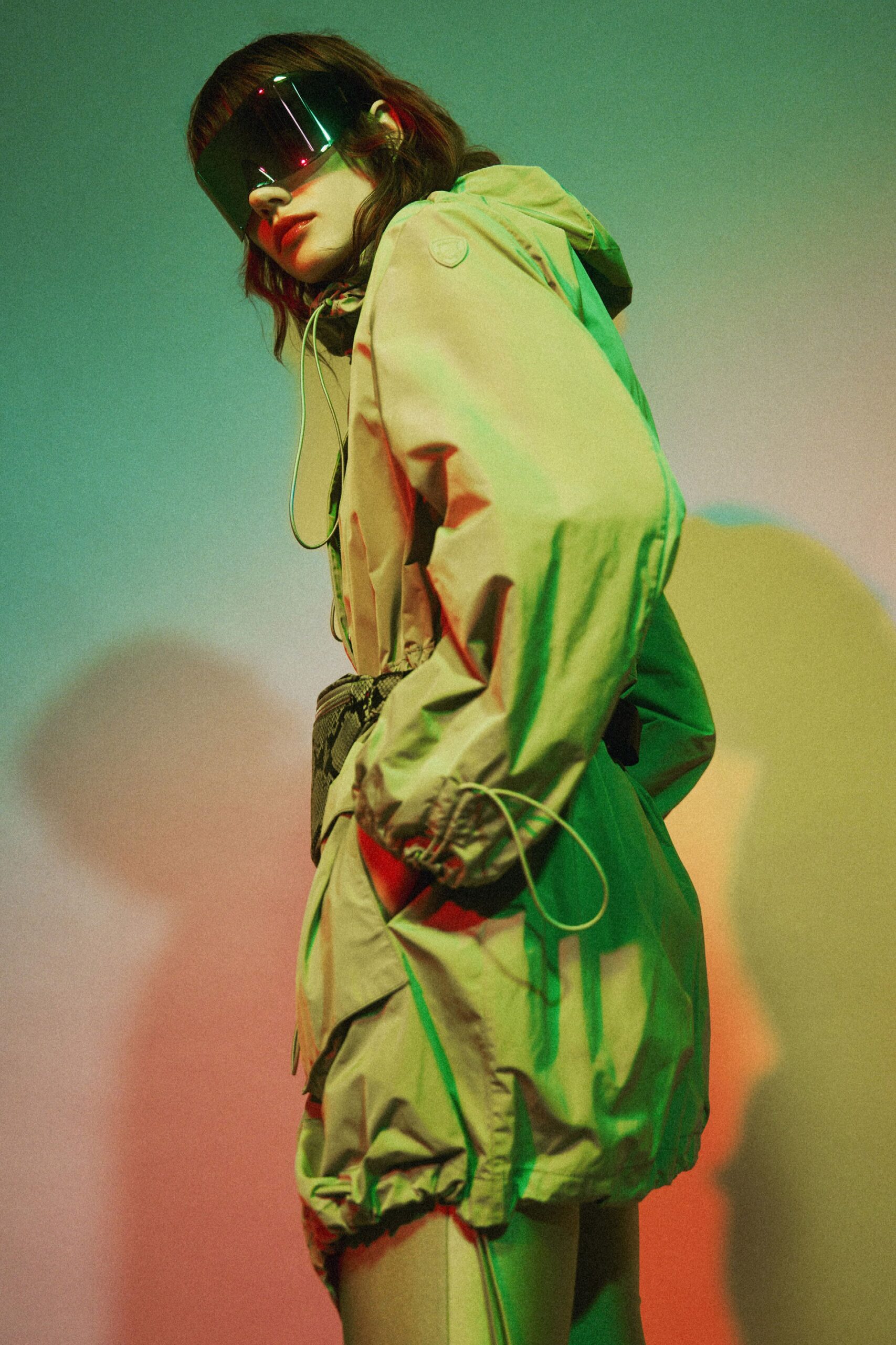 Oversized black shield lens catching pinprick red reflections. Pale sage nylon anorak, bungee cords dangling past the gathered cuff. Python-pattern belt bag strapped tight at the waist. Second-skin leggings with a raised side seam. Walking under a flyover after rain, every puddle throwing back neon.