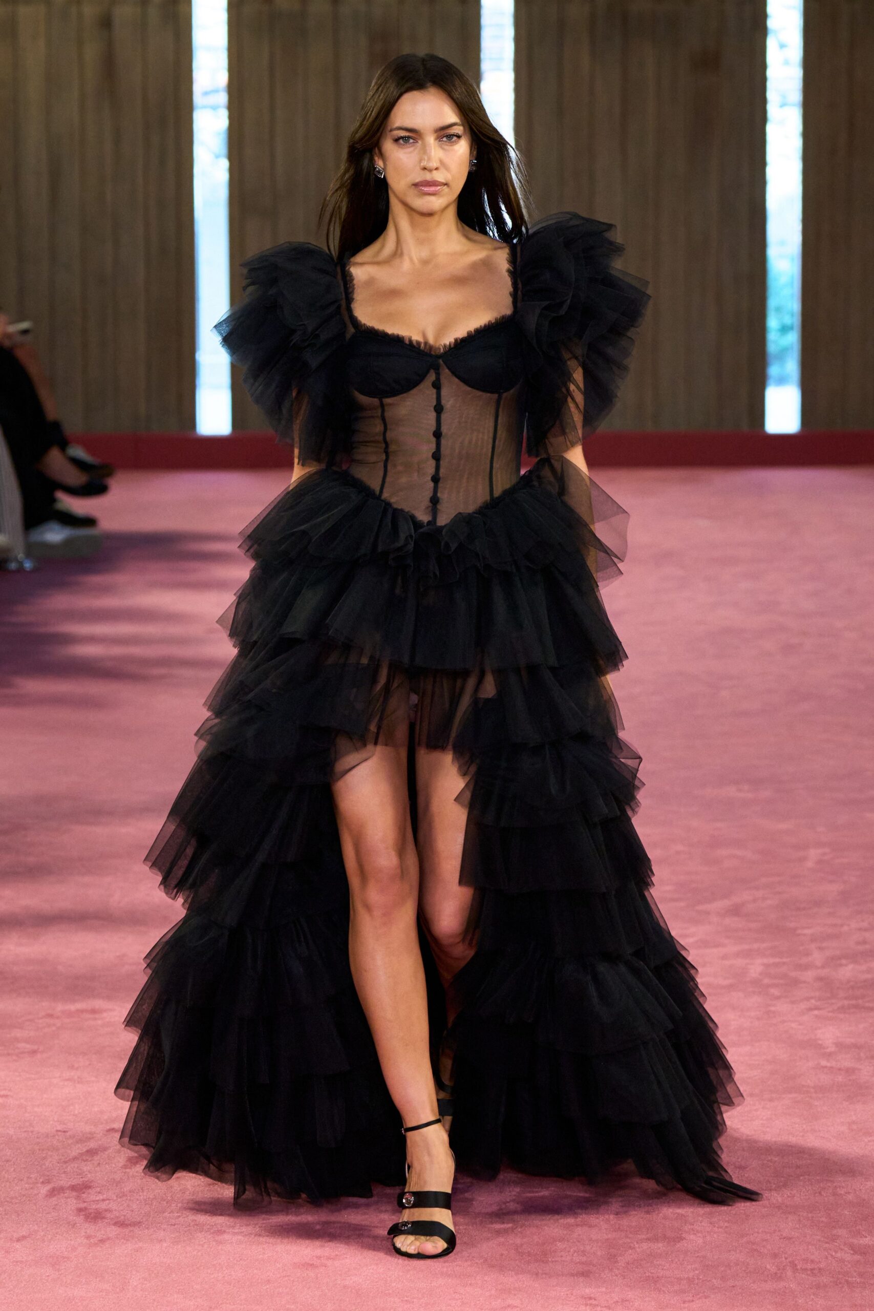 Black tulle dress with sheer corseted bodice, puffed sleeves and a tiered high-low train. Black satin mule sandals with two crystal-button straps. Walking across a hotel ballroom carpet after the orchestra has packed up.