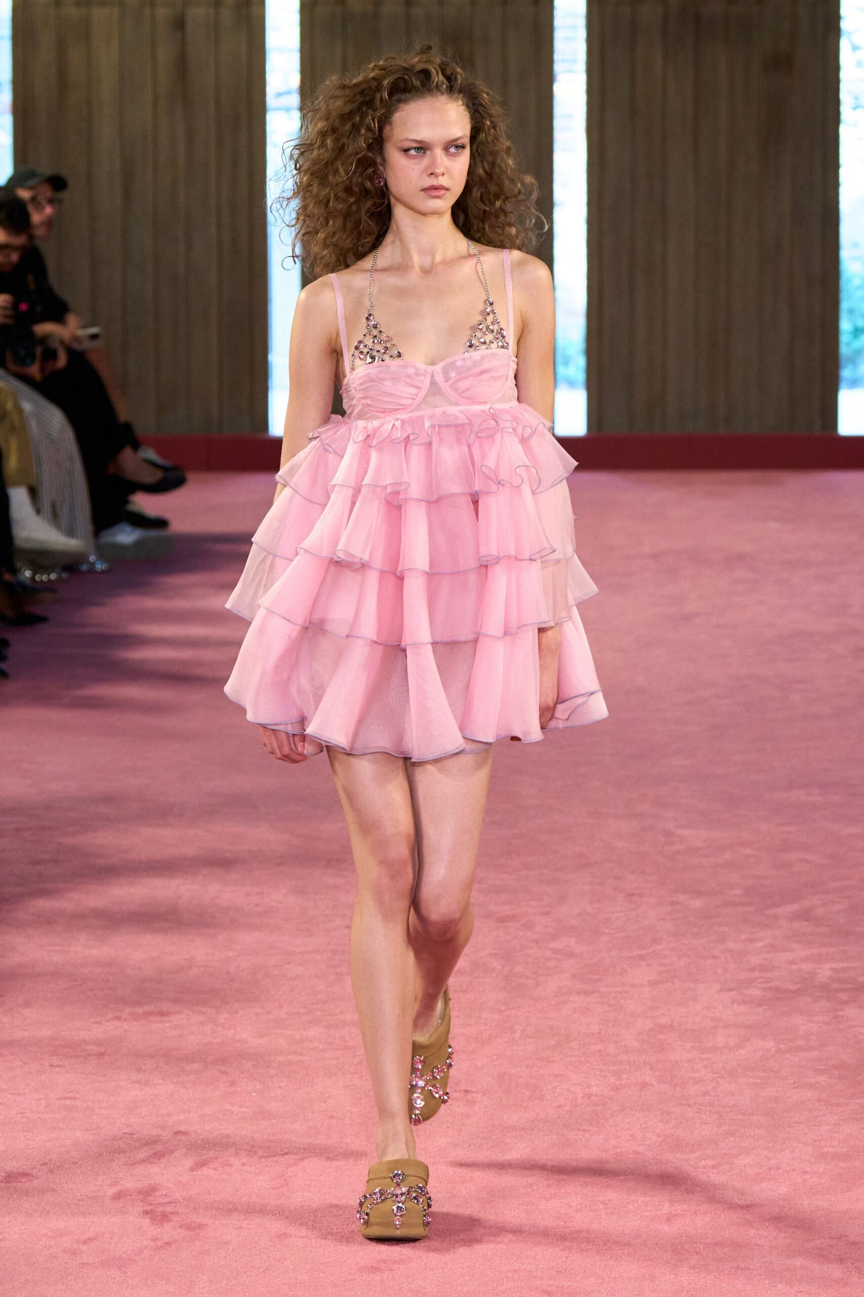 Pink organza babydoll dress tiered in floaty ruffles with grey picot edging. Crystal-linked halter triangles glinting above the ruched bust cups. Tan satin platform mules crossed with pink gem straps. Walking across hotel carpet toward a ballroom wedding that starts at noon.