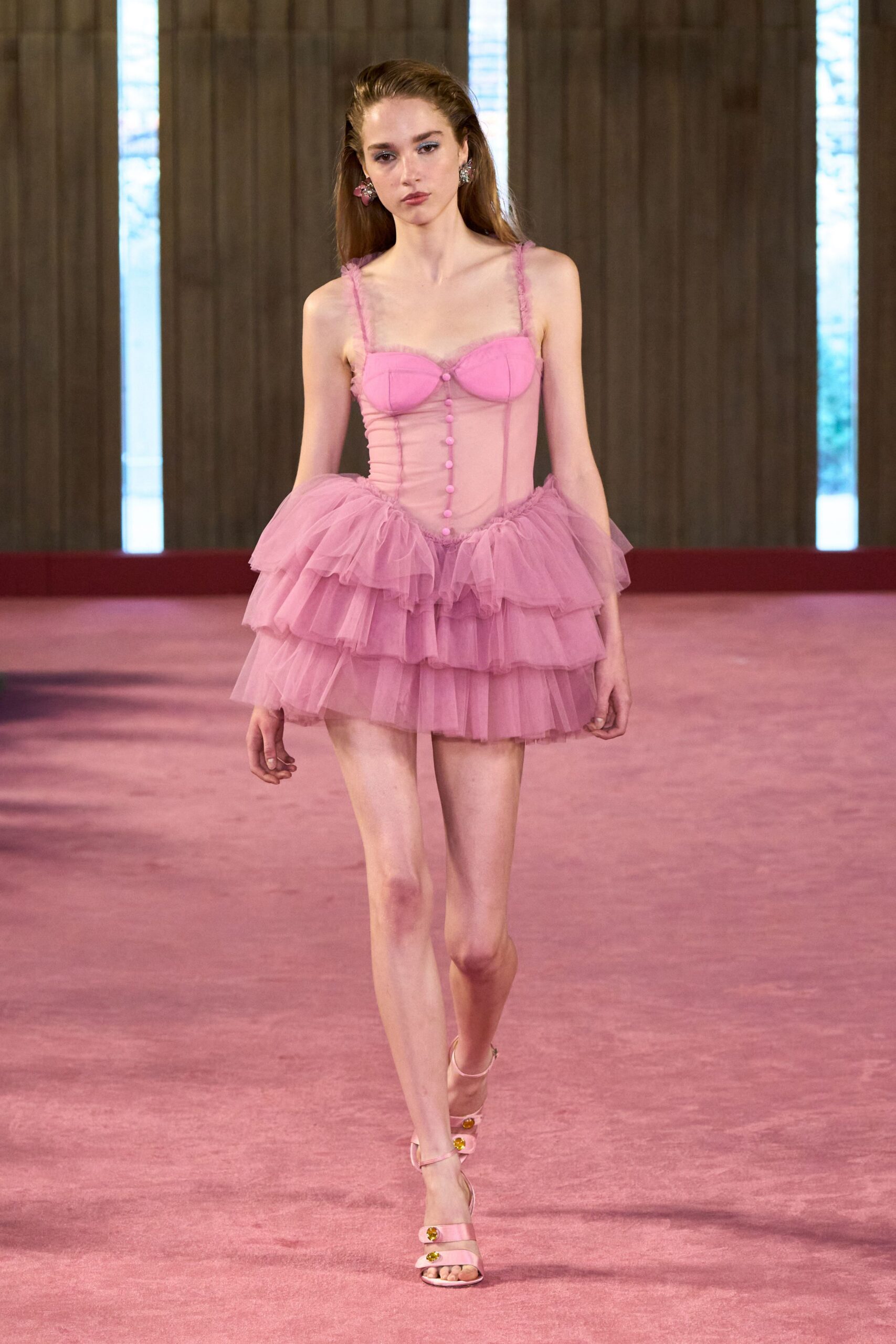 Mismatched blossom earrings, one pink and one silver. Ballet-pink mesh corset dress with molded cups and tulle straps. Tiered tulle mini flounce, waist ruffles breaking into soft volume. Pale pink satin sandals with jewel buttons on each strap. For cake in a hotel suite after the bridesmaids already left.