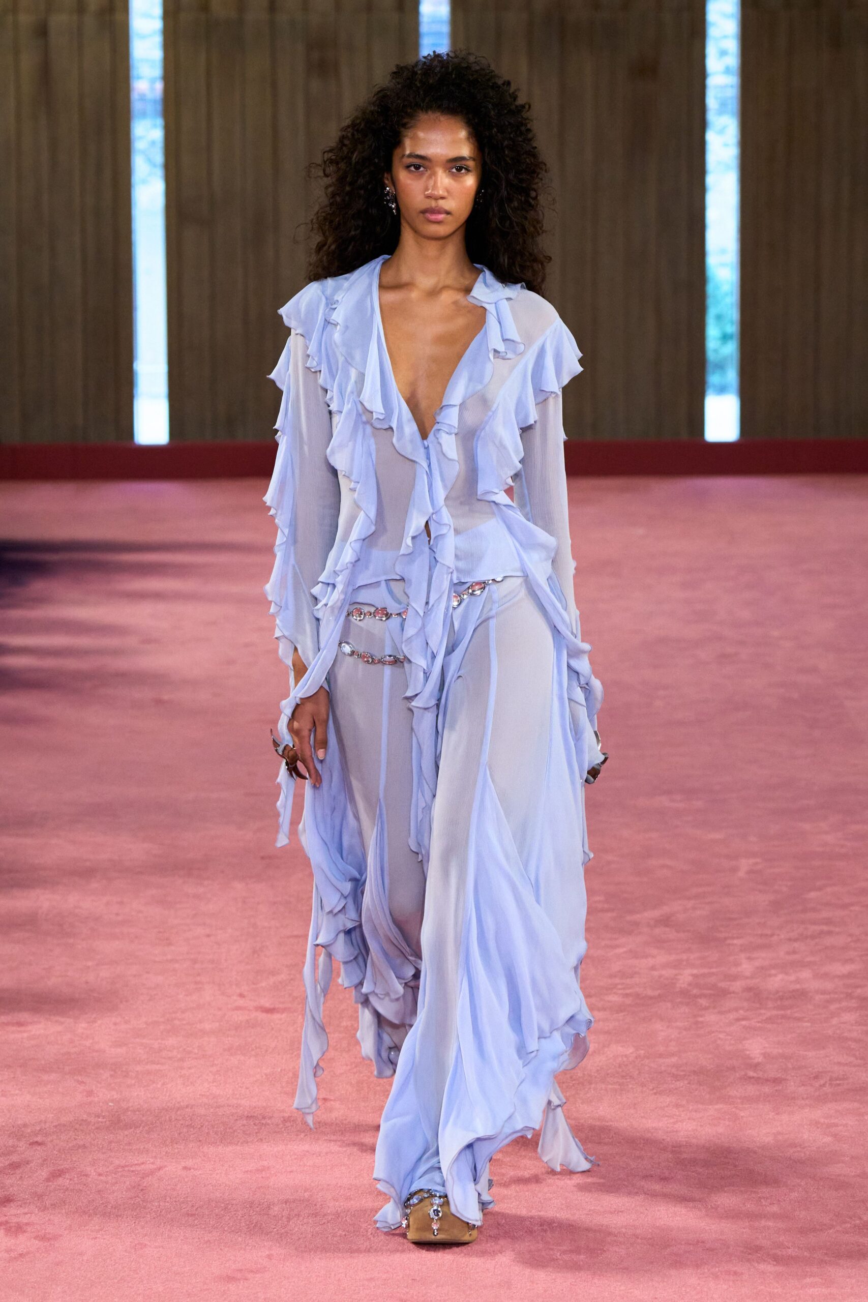 Powder-blue sheer georgette blouse with cascading lettuce-edge ruffles. Matching fluid trousers, split by two crystal chains at the hip. Nude satin mules with a dangling crystal drop at the toe. Walking through a hotel atrium before anyone else is awake.