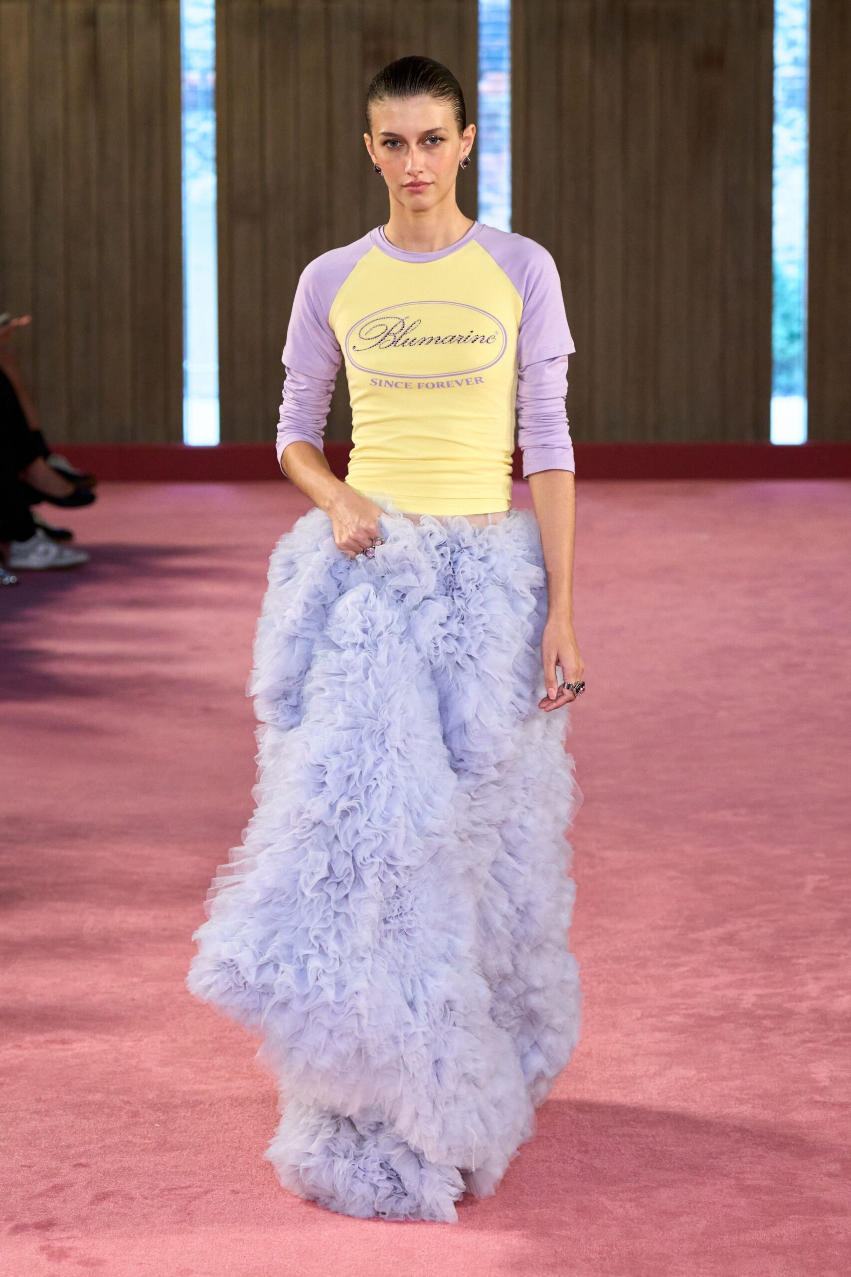 Butter yellow jersey raglan tee with lilac sleeves and printed oval logo. Powder-blue tulle skirt exploding in deep rosette ruffles from hip to floor. Small dark stud earrings. Chunky flower-shaped rings catching at her fingers. Hotel ballroom carpet at noon, still dressed for the cake reveal.