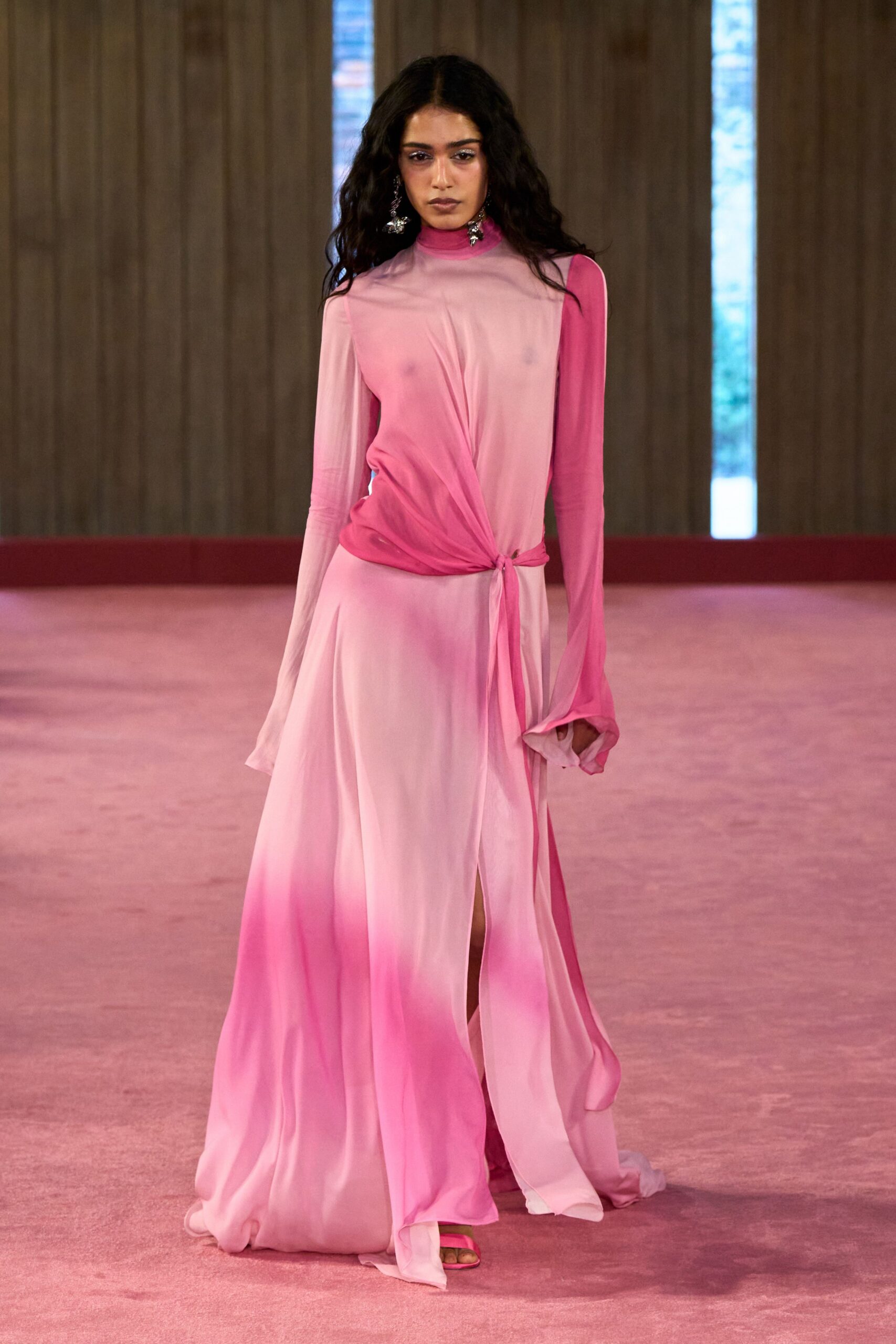 Oxidized silver drops with clustered charms brushing the jaw. Sheer blush jersey dress airbrushed deeper pink at throat and hem, twisted into a side knot. Hot-pink satin sandals peeking from the pooled front slit. Walking barefoot home through a hotel corridor after the wedding finally emptied.