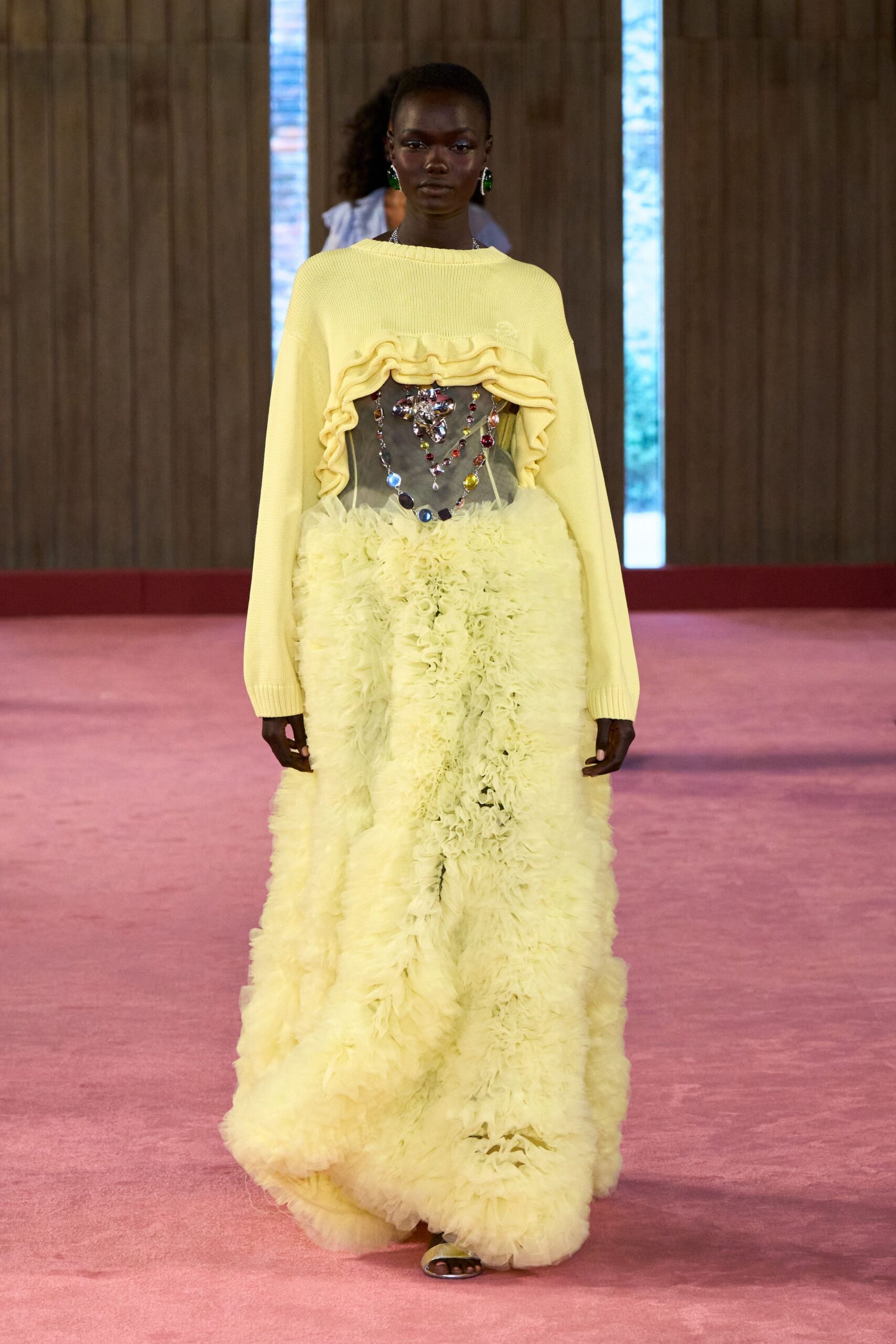Pale citron ribbed knit, cropped in a deep ruffle above the bust. Sheer corset strung with oversized jewel drops across the front. Floor-length tulle skirt in dense whipped ruffles. Pale satin toe-band sandals. Large green oval drops. Afternoon wedding in a glasshouse, someone carrying the cake too carefully.
