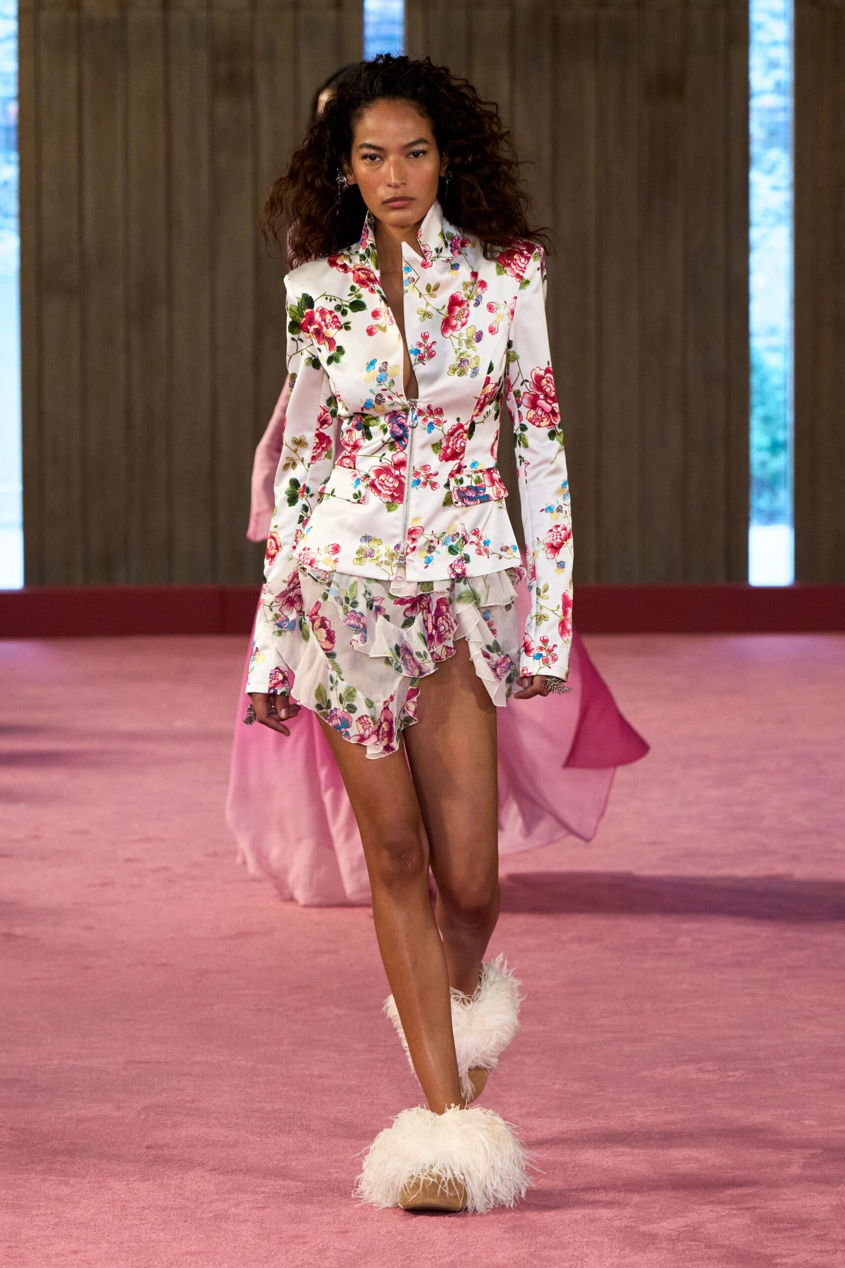 Ivory satin jacket printed with cabbage roses, stand collar framing a deep slit. Sheer floral miniskirt with asymmetric chiffon ruffles kicking from the hip. Tan mules swallowed by shaggy white marabou. Garden wedding reception, someone dragged the speakers onto the lawn.