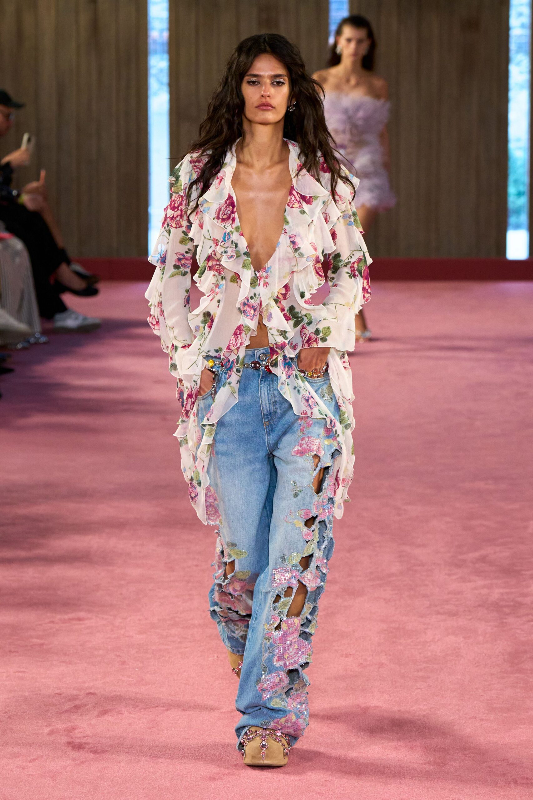 Sheer ivory georgette blouse, rose print tumbling into deep ruffled fronts. Low slung jewel belt with one dark oval at center. Pale denim slashed open and appliqued with sequined pink blossoms. Nude satin mules crusted with pink stones at the square toe. Walking back from a beach wedding after kicking off the formal plan.