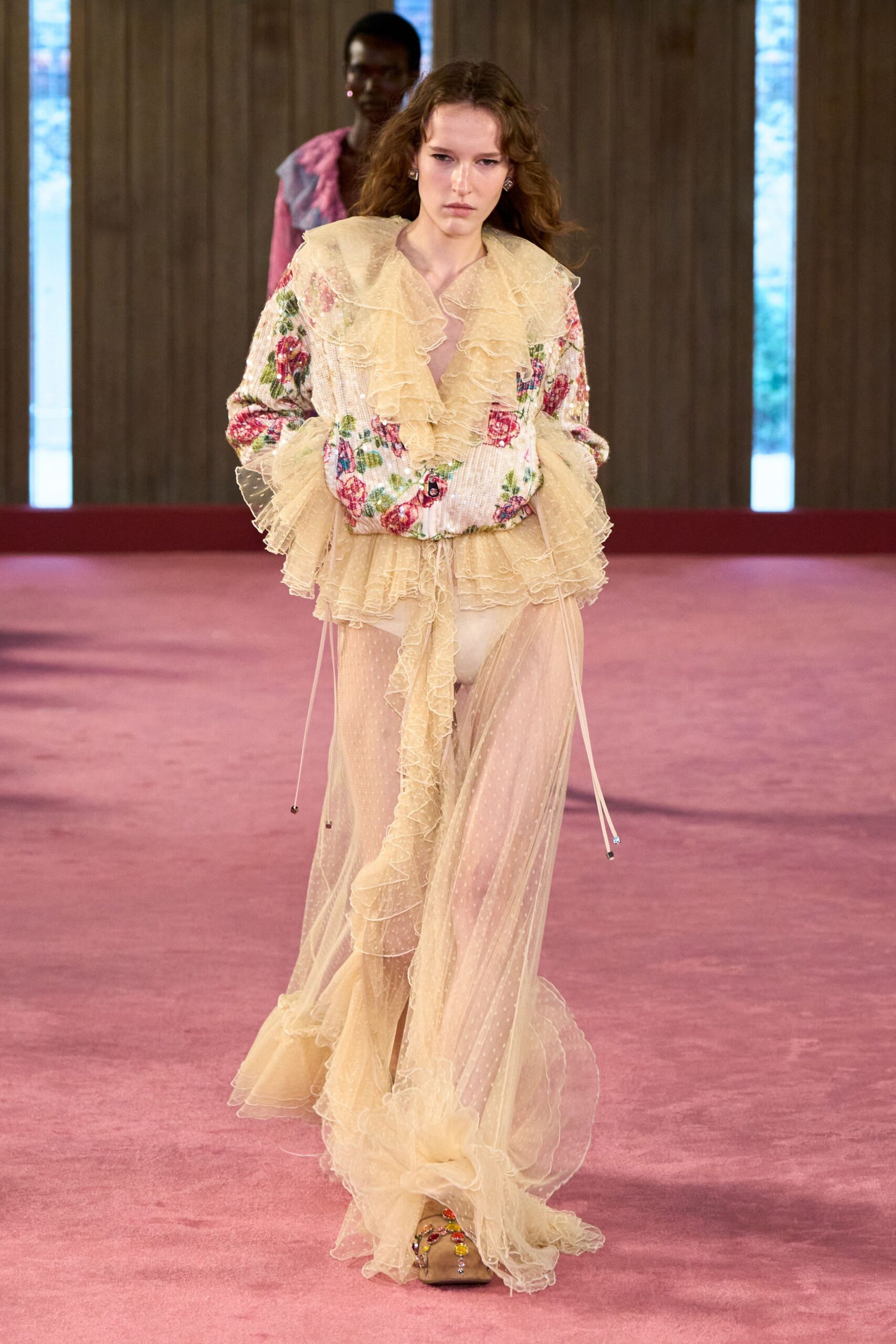Buttercream net blouse with a plunging ruffle cascade and sequin floral sleeves. Sheer dotted tulle trousers, split open under a long frilled panel. Nude briefs visible beneath. Square-toe mules crusted with cabochon jewels across the vamp. Walking back through a faded hotel ballroom after the wedding band packed up.