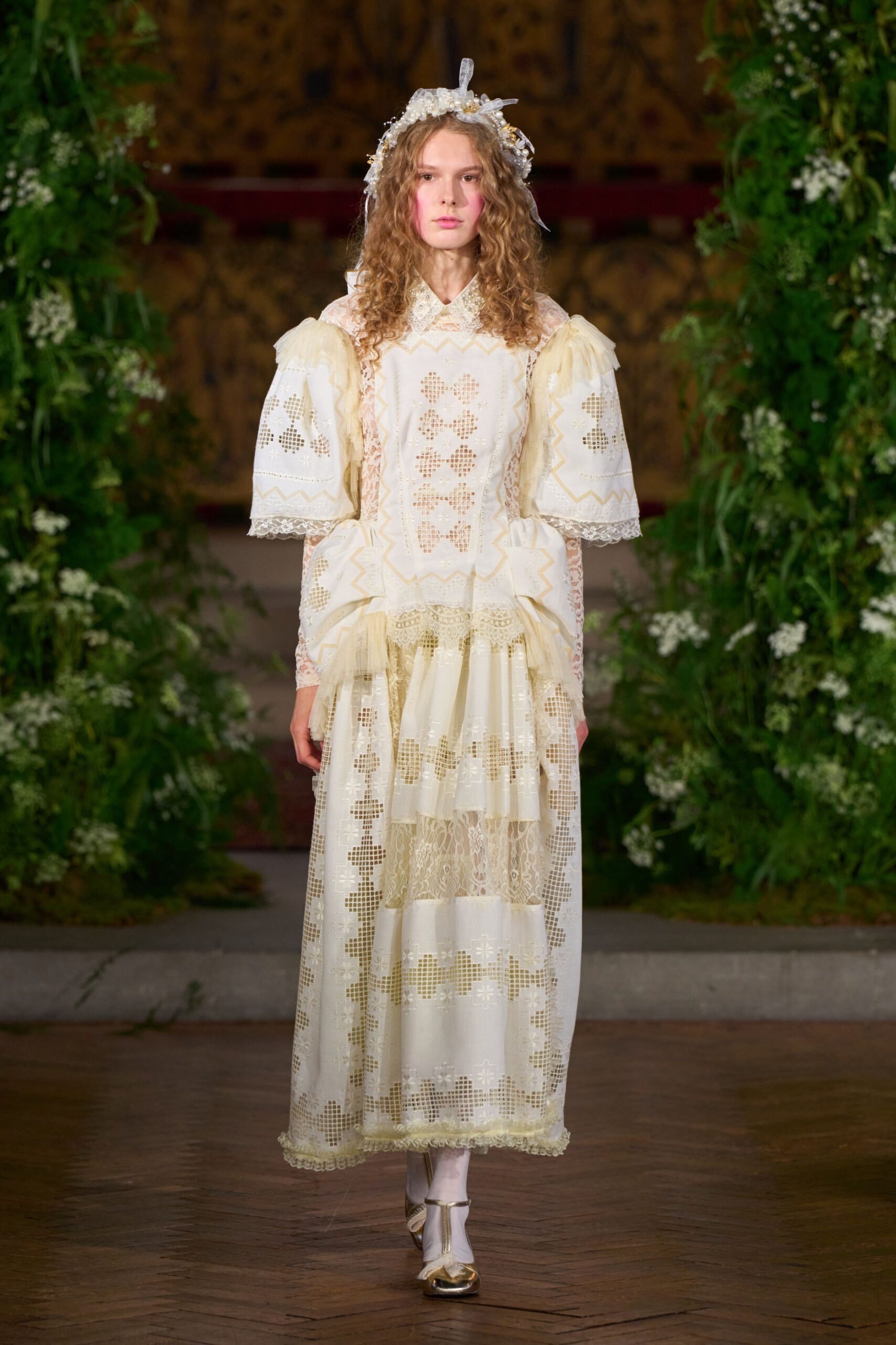 Pearled floral crown with wired petals and loose ribbon ends. Ivory cutwork dress with zigzag inserts, puffed sleeves and lace bands. White stockings with a centered gold seam. Gold cap-toe Mary Janes, bow-tied and slightly blunt. Walking through a walled garden after the wedding cake has been cut.