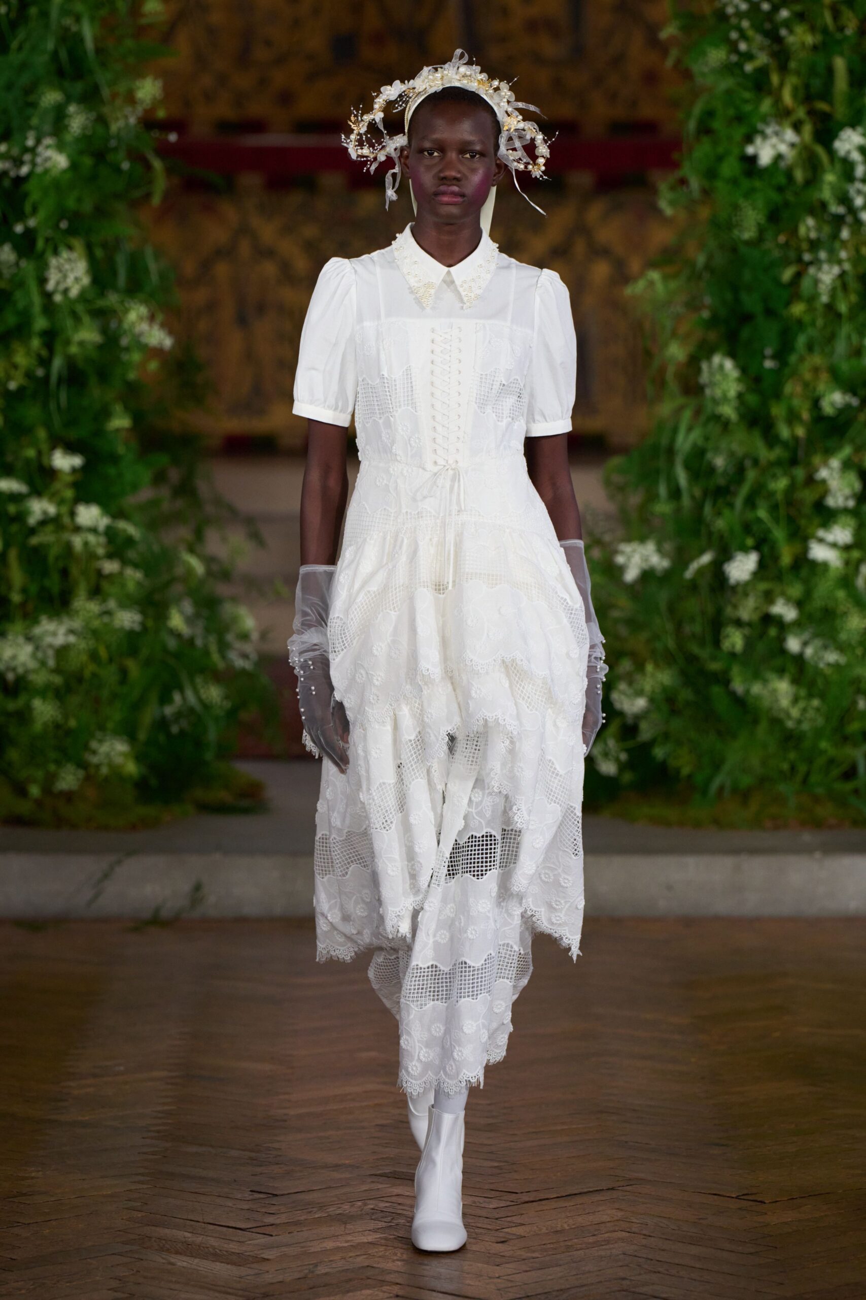 Pearled wire halo with sheer ribbon streamers and tiny silk blossoms. White lace-patchwork dress, corset lacing and beaded collar over puff sleeves. Sheer tulle gloves dotted with dangling pearl drops. White leather ankle boots with a plain rounded toe. Crossing the orangery after the ceremony, petals stuck to the parquet.