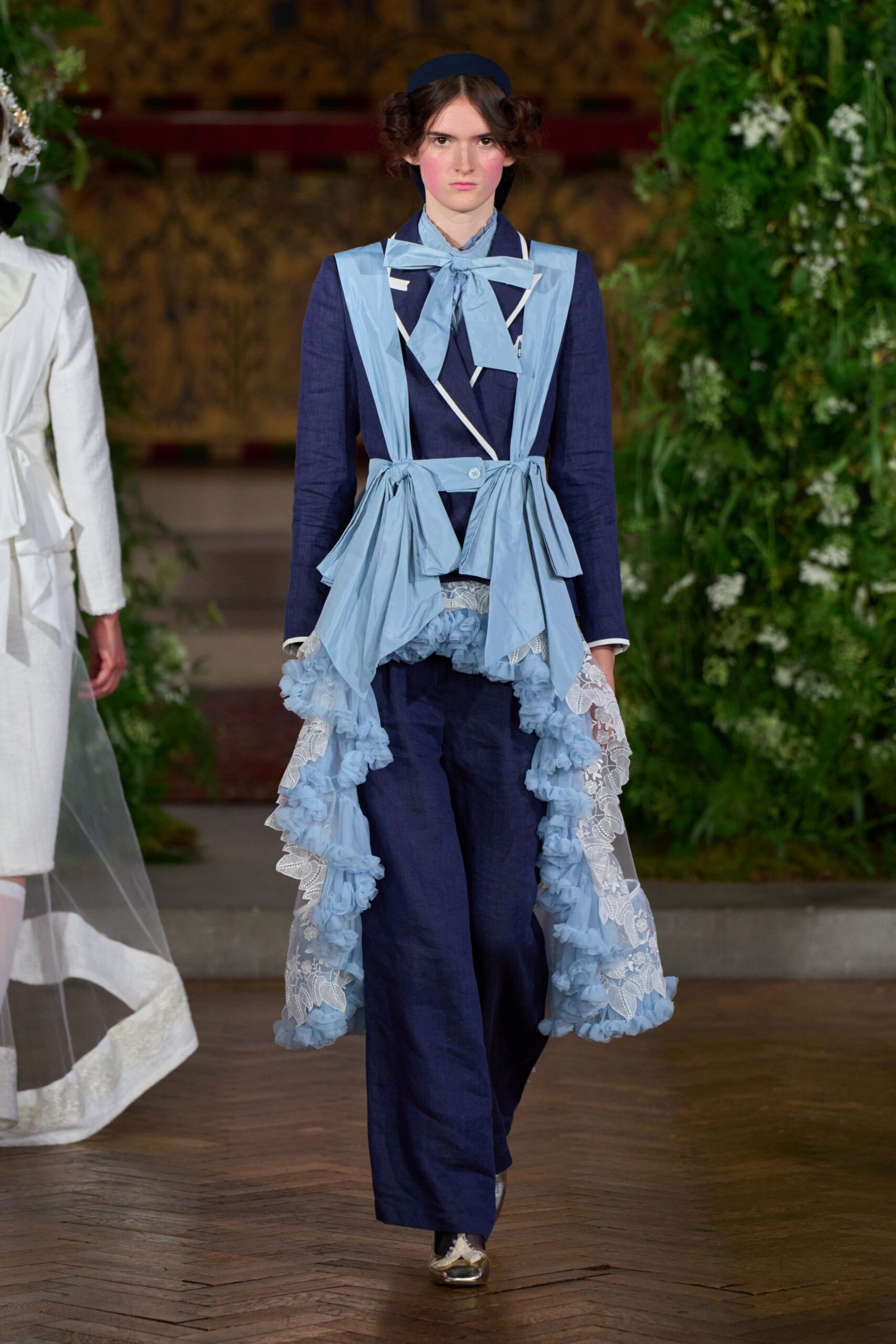 Navy headband behind looped side buns. Ink linen jacket with white piping and a pale blue neck bow. Powder-blue panels tied at the waist, trailing lace and ruffled chiffon. Matching wide trousers in the same slubby linen. Silver flats with a frayed cream bow. Crossing a manor hallway after the wedding cake has already been cut.