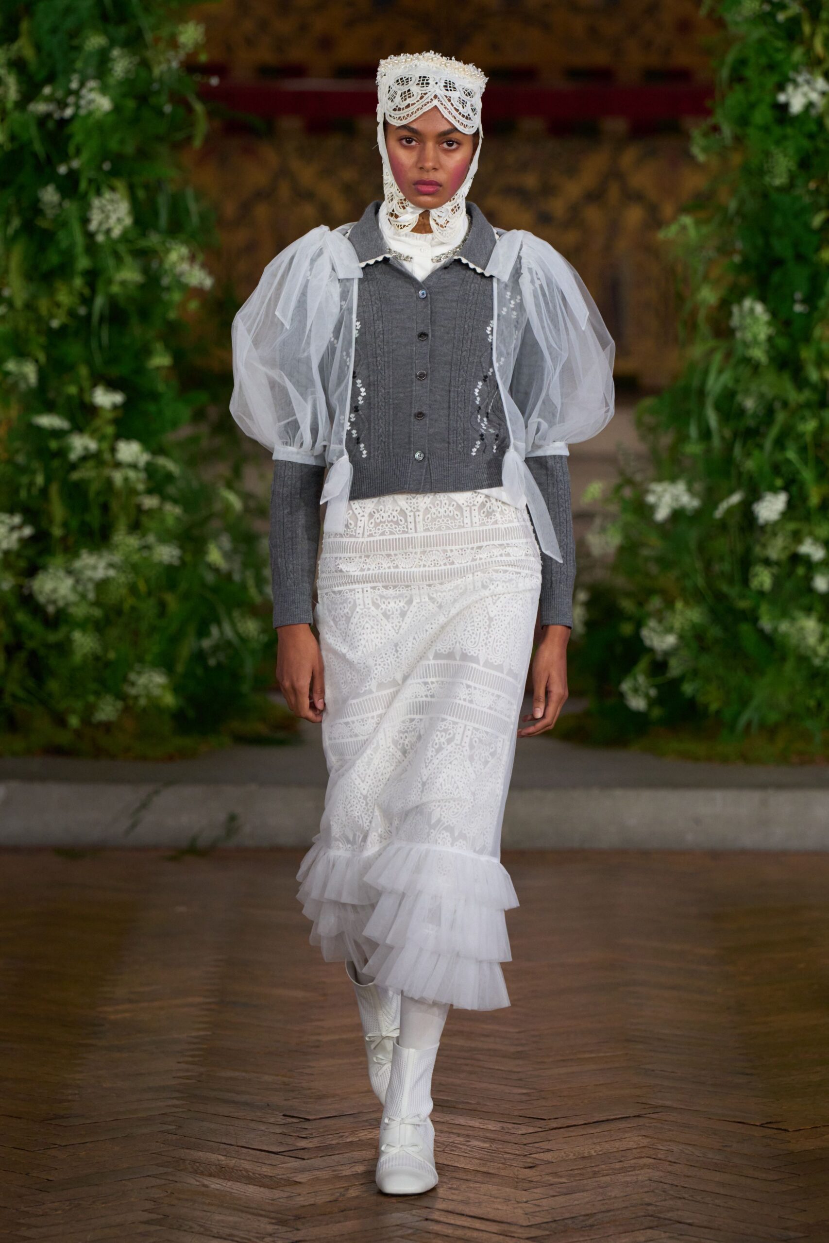 White cutwork lace bonnet framing the face and throat. Grey knit cardigan, pin-tucked front under sheer puffed organza sleeves. White broderie anglaise column skirt with tiered tulle flounce. White knit boots, tiny bows climbing the front. Crossing a manor hallway after the wedding cake has been cleared.