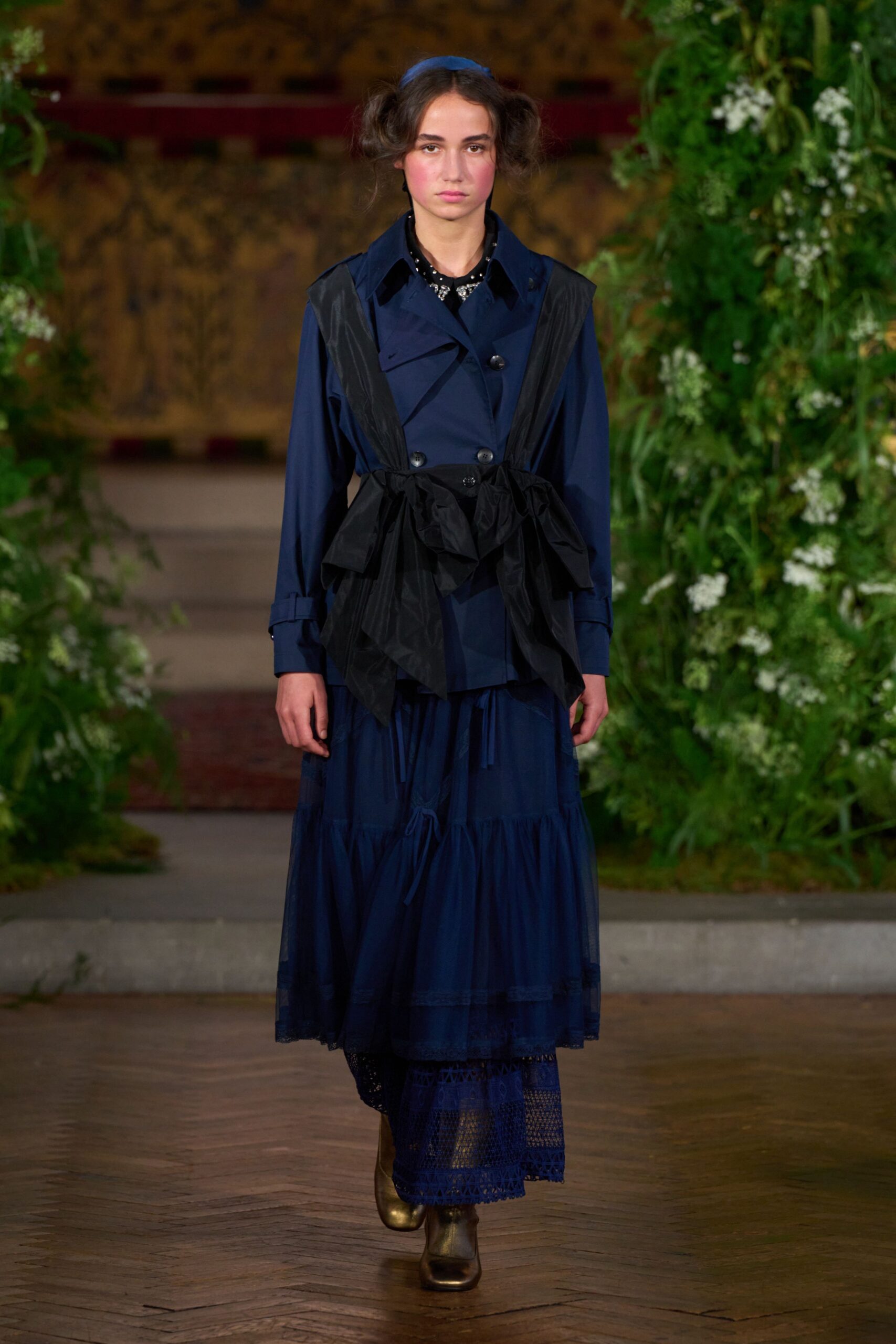 Cobalt satin headband above puffed pigtail buns. Navy double-breasted trench, cinched with a crinkled black overskirt panel. Midnight tiered dress with sheer lace peeking below the hem. Bronze square-toe boots. Walking through a walled garden after rain, hems taking the damp.