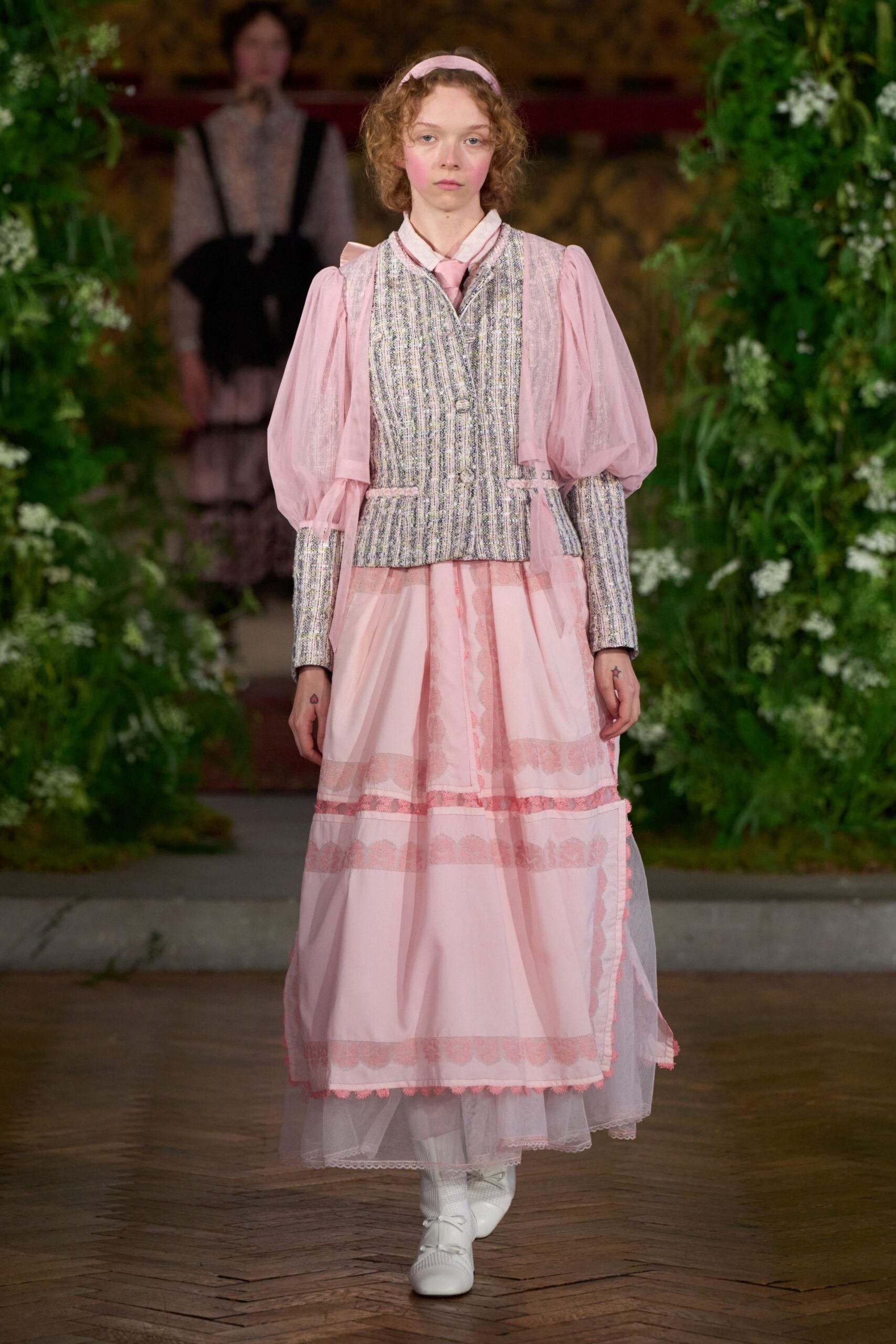 Pale pink satin headband over loose corkscrew curls. Powder pink dress with sheer puff sleeves and lace-banded tiers. Pastel bouclé vest, two fabric-covered buttons and pink trim tabs. White lace-up boots with ribbed sock tops peeking above. Crossing the orangery after a children's recital, still carrying the blush.