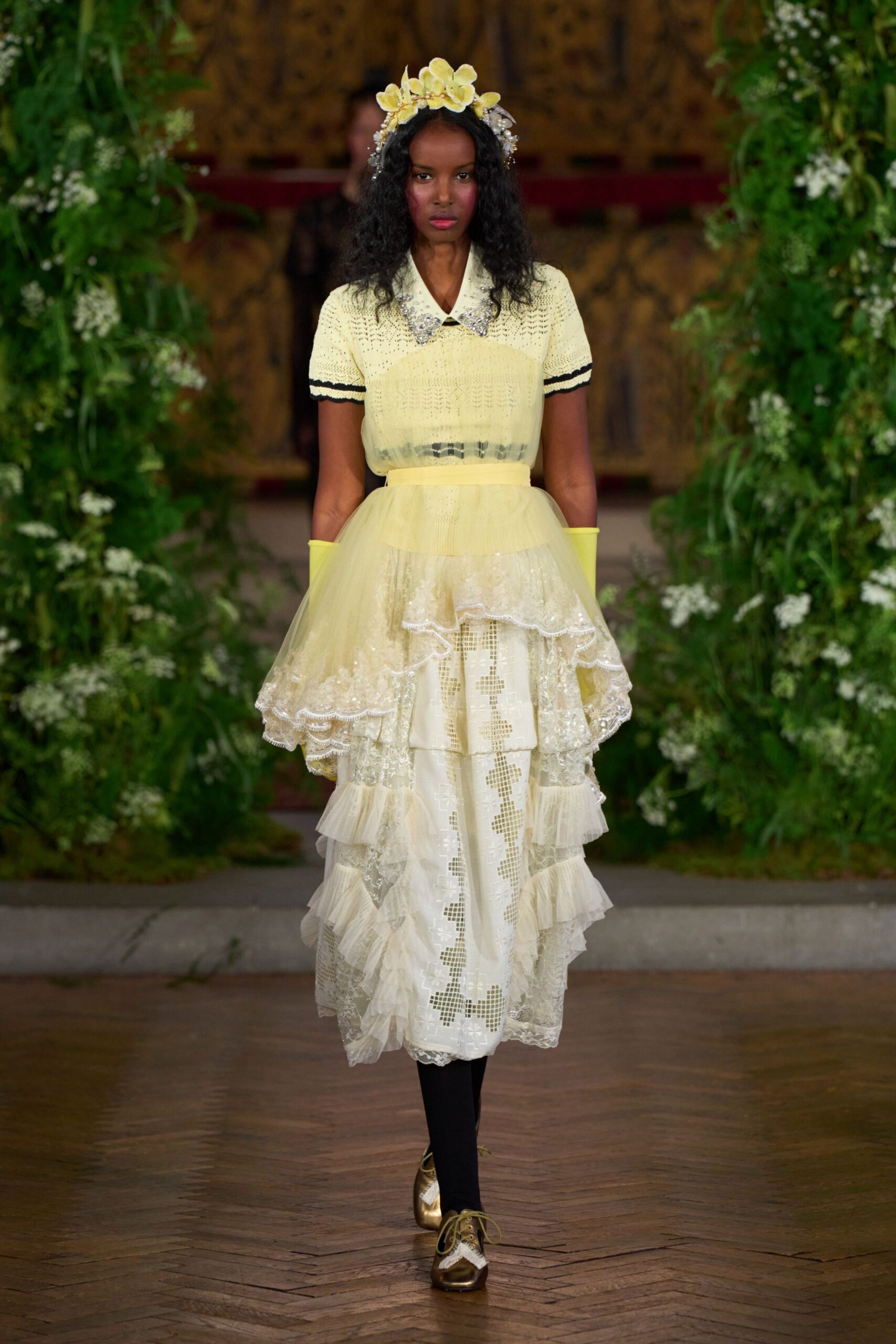 Butter-yellow orchids and pearl sprigs perched in the hair. Pale lemon open-knit top with a rhinestone-spiked collar. Chartreuse opera gloves. Tiered ivory lace skirt under a sheer yellow tulle peplum. Opaque black tights. Gold leather oxfords with white brogue inserts. Garden wedding, rain held off but the grass is still wet.