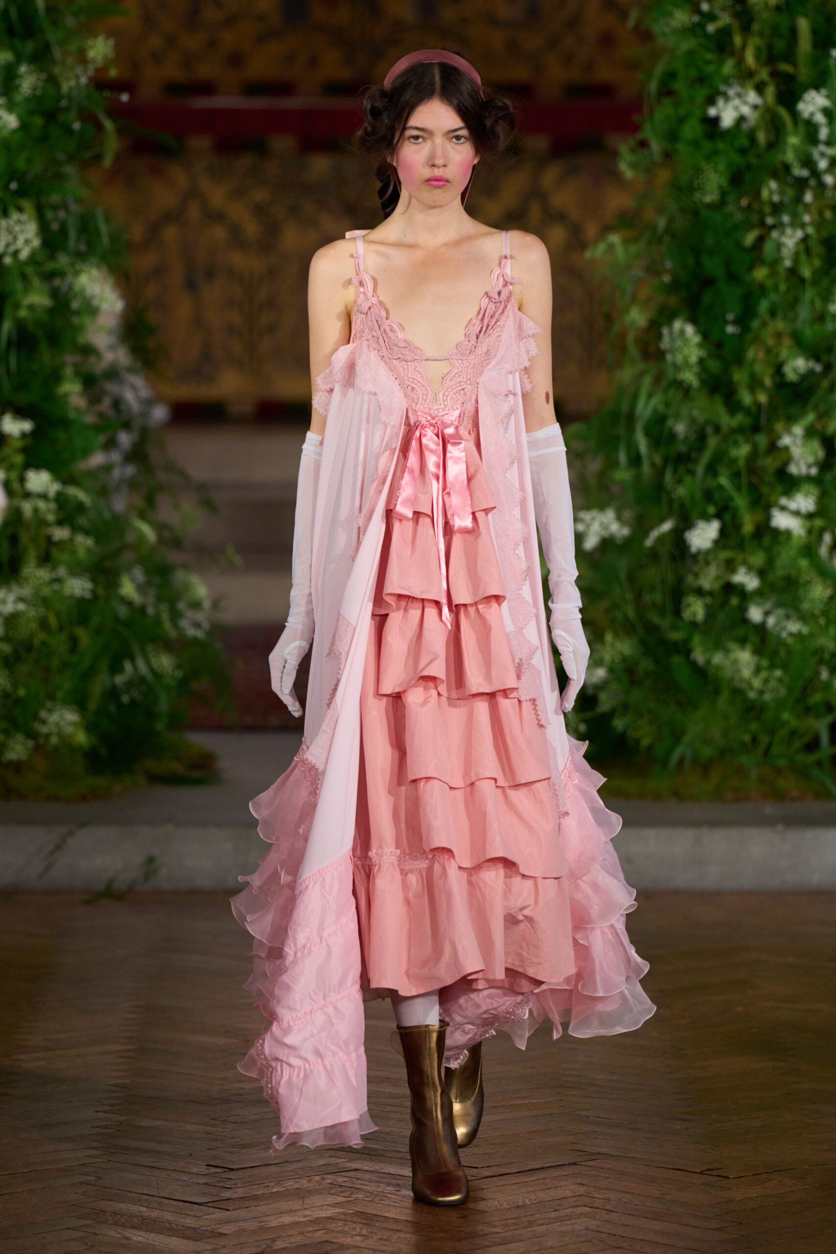 Wide rose satin headband over coiled side buns. Blush slip dress with scalloped lace plunge and a glossy bow. Sheer pink overskirt edged in lace, organza ruffles pooling behind. Elbow-length white mesh gloves. Bronze ankle boots, square at the toe. Walking across the conservatory after the wedding guests have drifted outside.