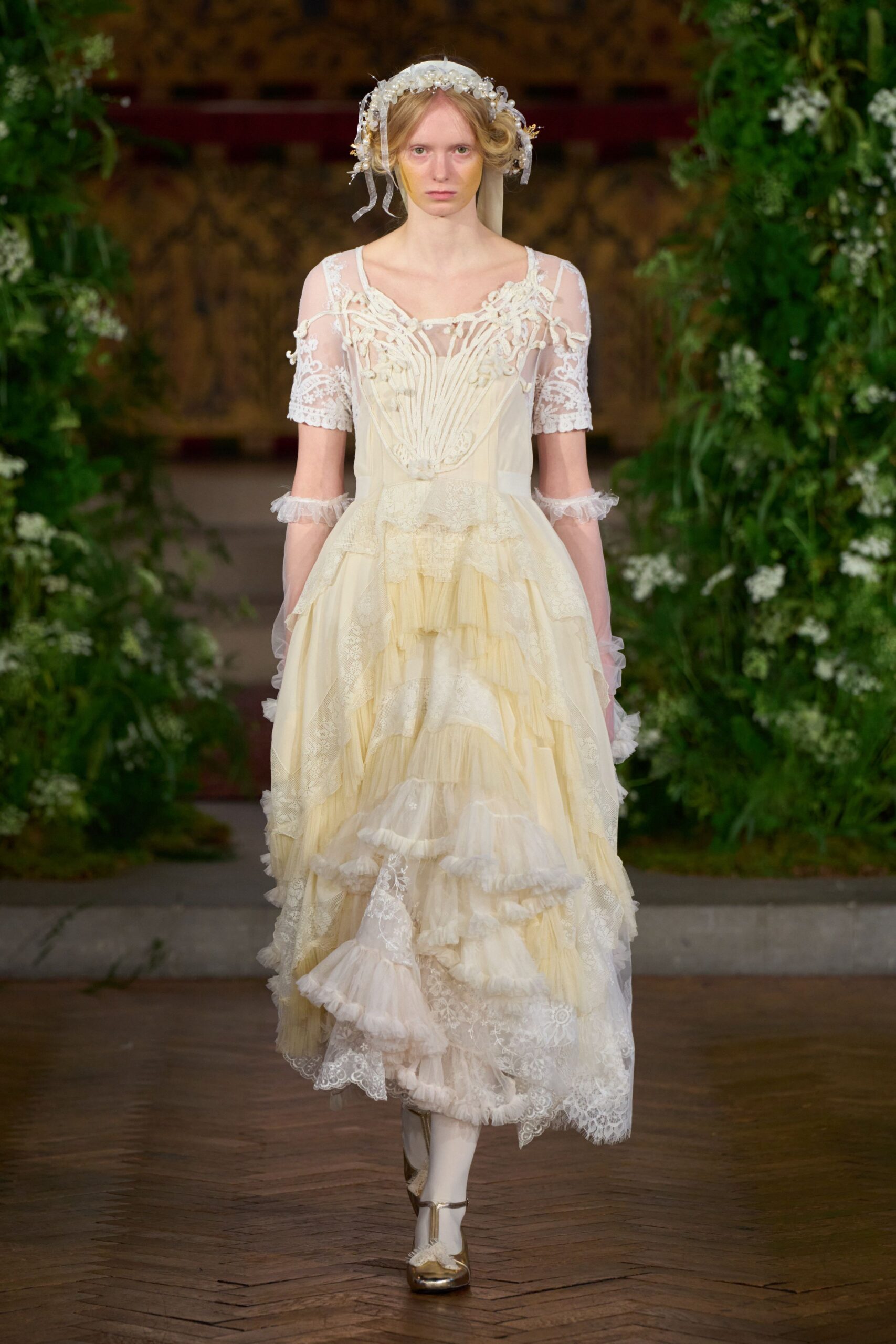 Pearled floral halo with ribbon loops and a trailing tulle veil. Ivory tulle dress with lace sleeves, corded shell bodice and uneven ruffled tiers. Sheer frills circling the forearms. Opaque white stockings. Gold Mary Jane heels with tiny bows at the toe. Crossing a damp lawn after the church bells have already stopped.