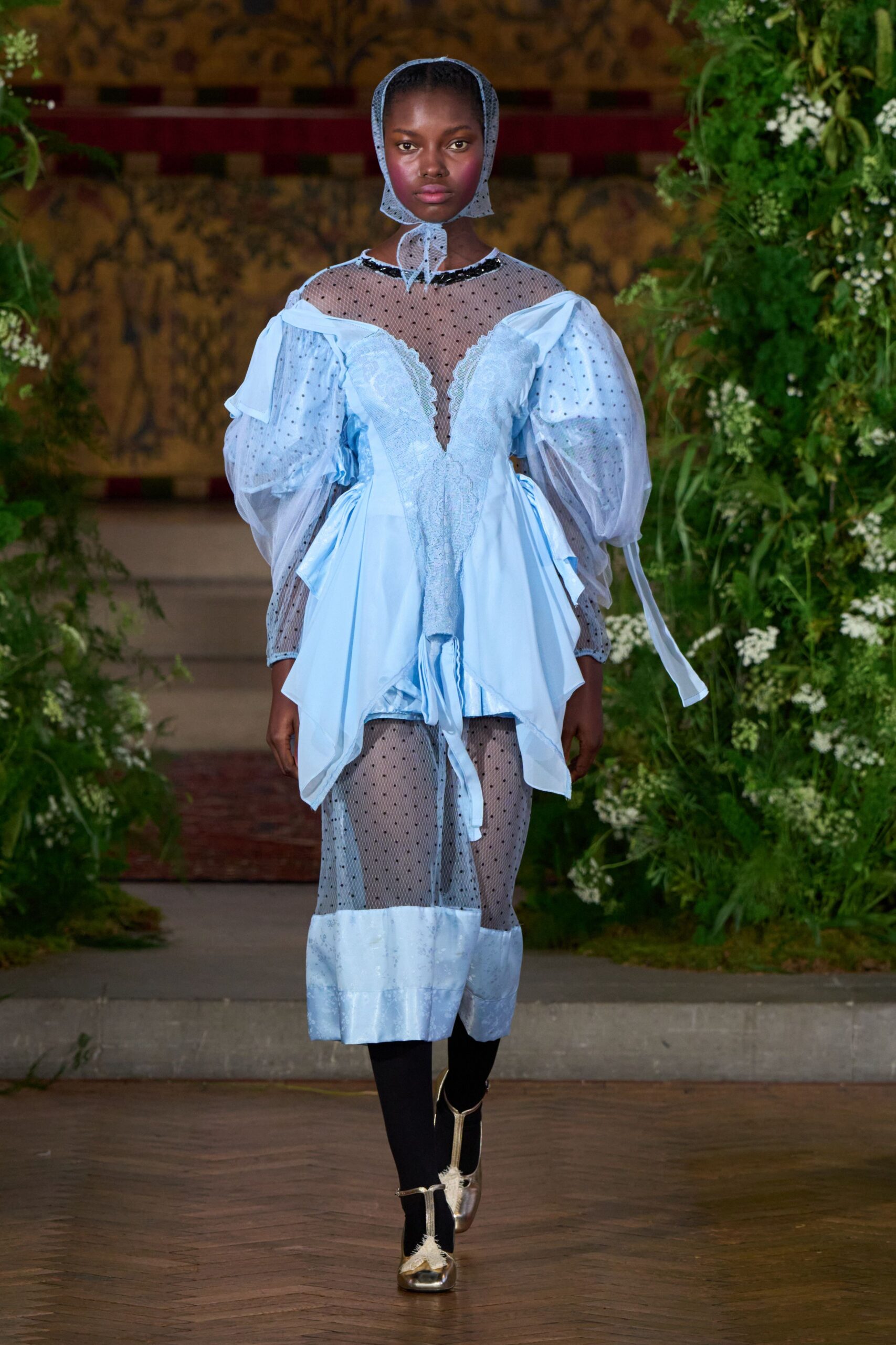 Powder-blue dotted tulle tied snugly under the chin. Ice-blue satin dress with sheer flocked mesh yoke and drooped puff sleeves. Matching dotted mesh stockings, satin cuffs grazing the calf. Black sock boots with silver shoes strapped over them. Crossing a formal garden after rain, hem already drinking from the path.