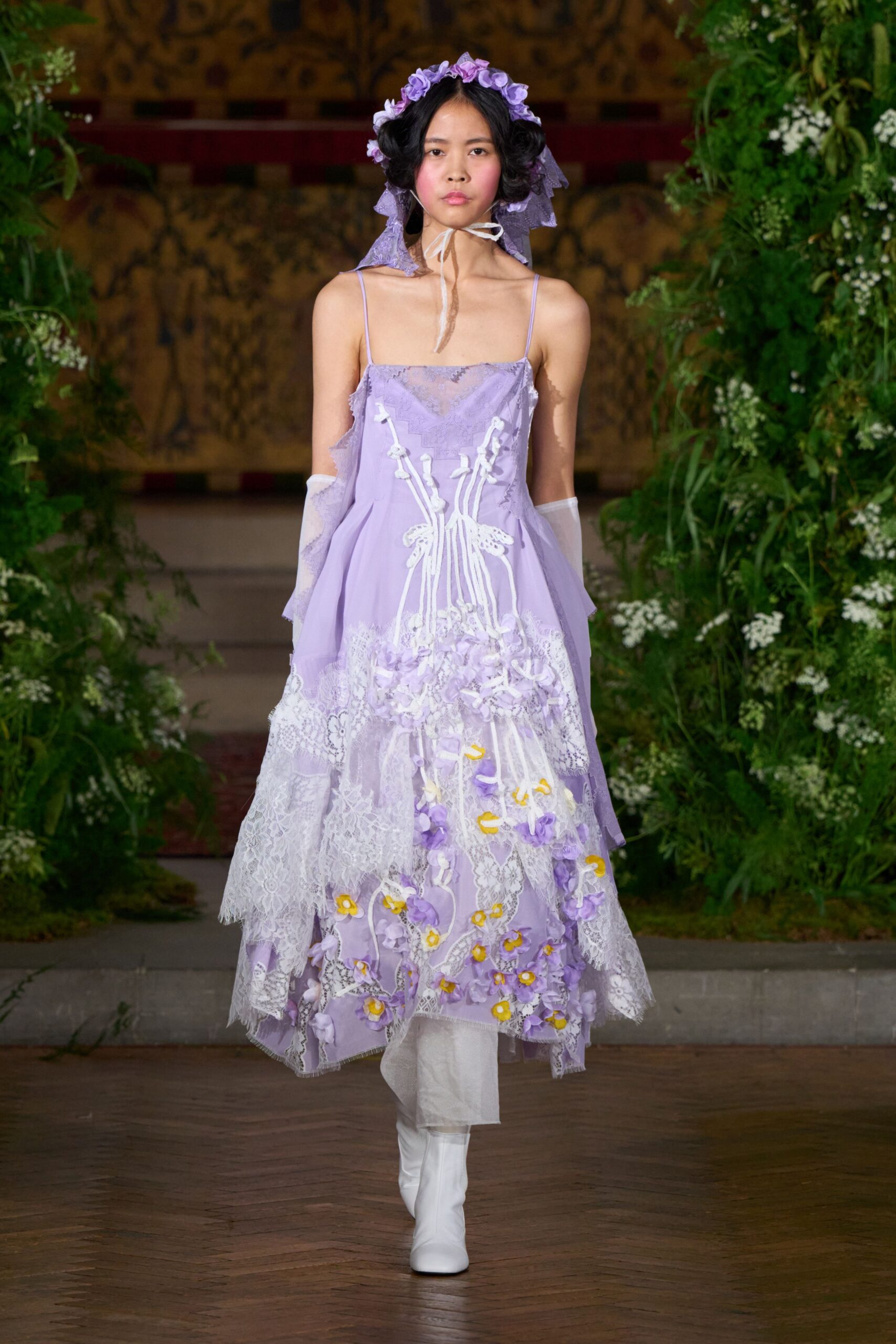 Lavender lace bonnet circled with lilac rosettes, tied under the chin. Lavender slip dress patched with torn lace and wired floral appliques spilling downward. Sheer white opera gloves. White knee boots, squared toes. Crossing the orangery after the garden wedding, petals stuck to the hem.