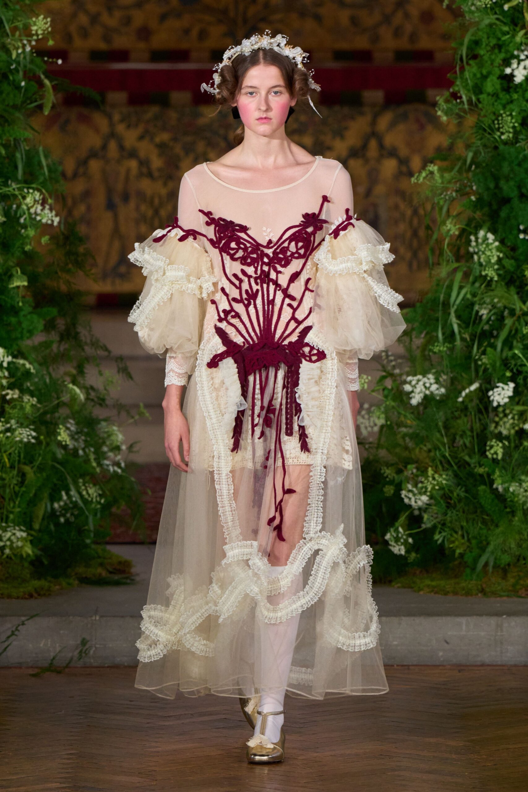 Crystal blossoms wired into the pinned coils. Sheer ivory tulle dress with dropped puff sleeves and ruffled lace bands. Burgundy corded vinework climbing the bodice, one tendril hanging past the hem. Pale pink stockings. Gold flats with ribbon ties. Crossing the manor ballroom after midnight, petals ground into the floorboards.